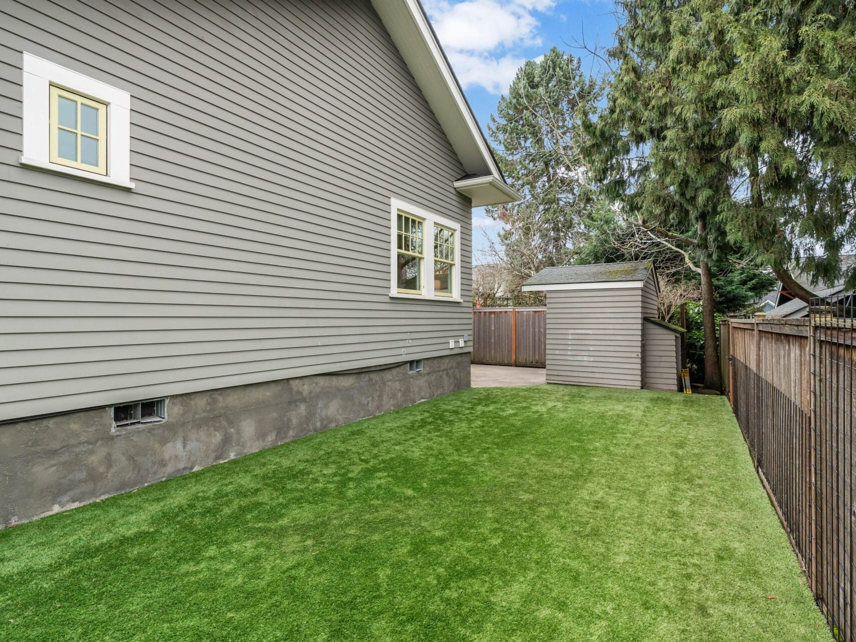 Side view of a modern house with gray siding, white-framed windows, and a neatly maintained lawn. A small shed is visible in the background next to a wooden fence, set against a backdrop of tall trees under a blue sky.