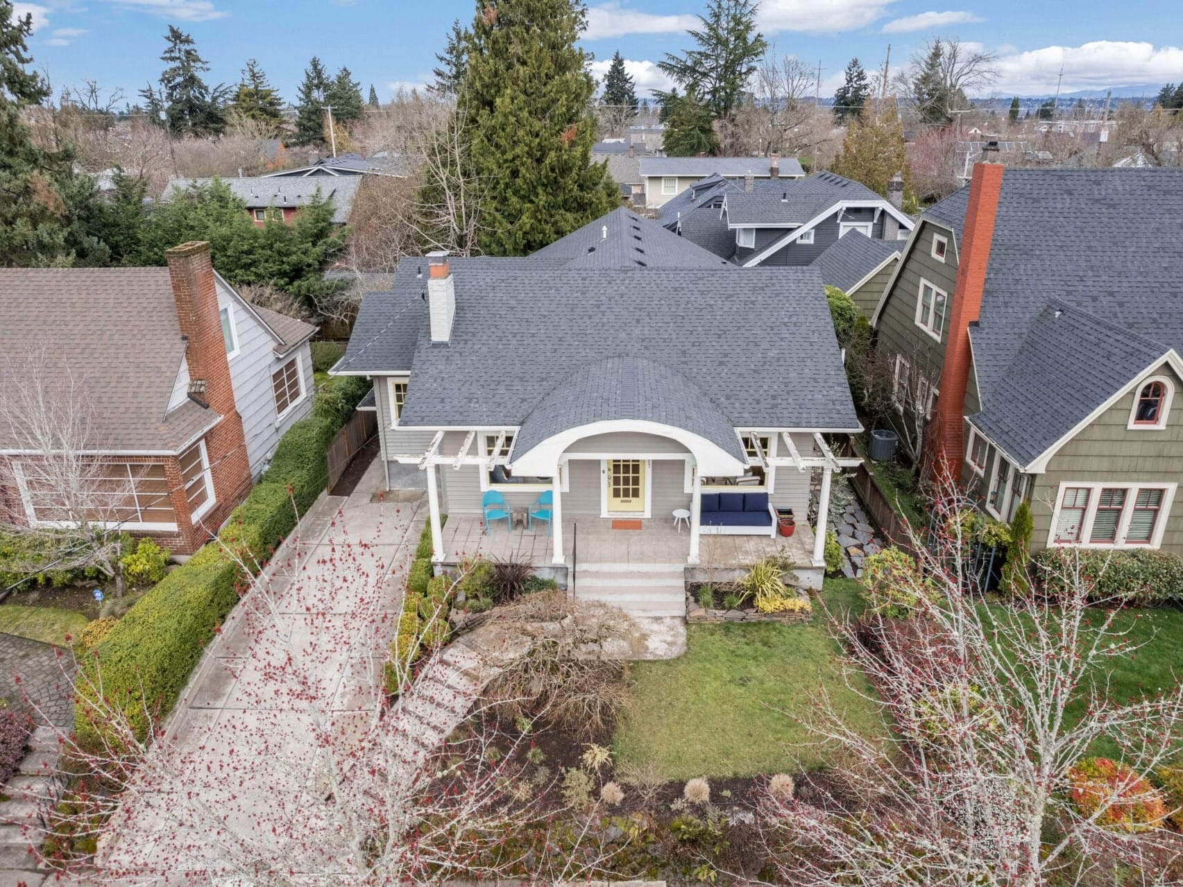 Aerial view of a suburban neighborhood featuring a charming house with a gray roof, front porch, and driveway. The house is surrounded by trees and neatly landscaped gardens, with neighboring houses visible on either side.