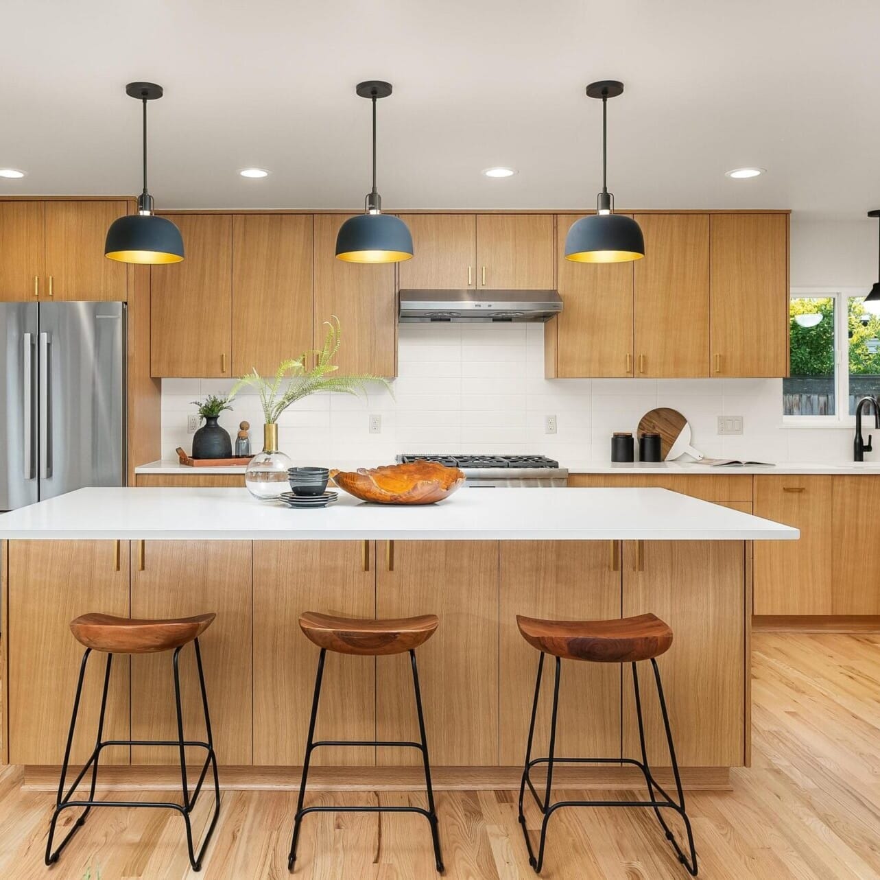 Modern kitchen with light wood cabinetry, a central island featuring a white countertop, and three wooden barstools. Overhead, black pendant lights delicately illuminate the space. Stainless steel appliances and decorative items complete this warm, inviting jewel in Portland real estate.