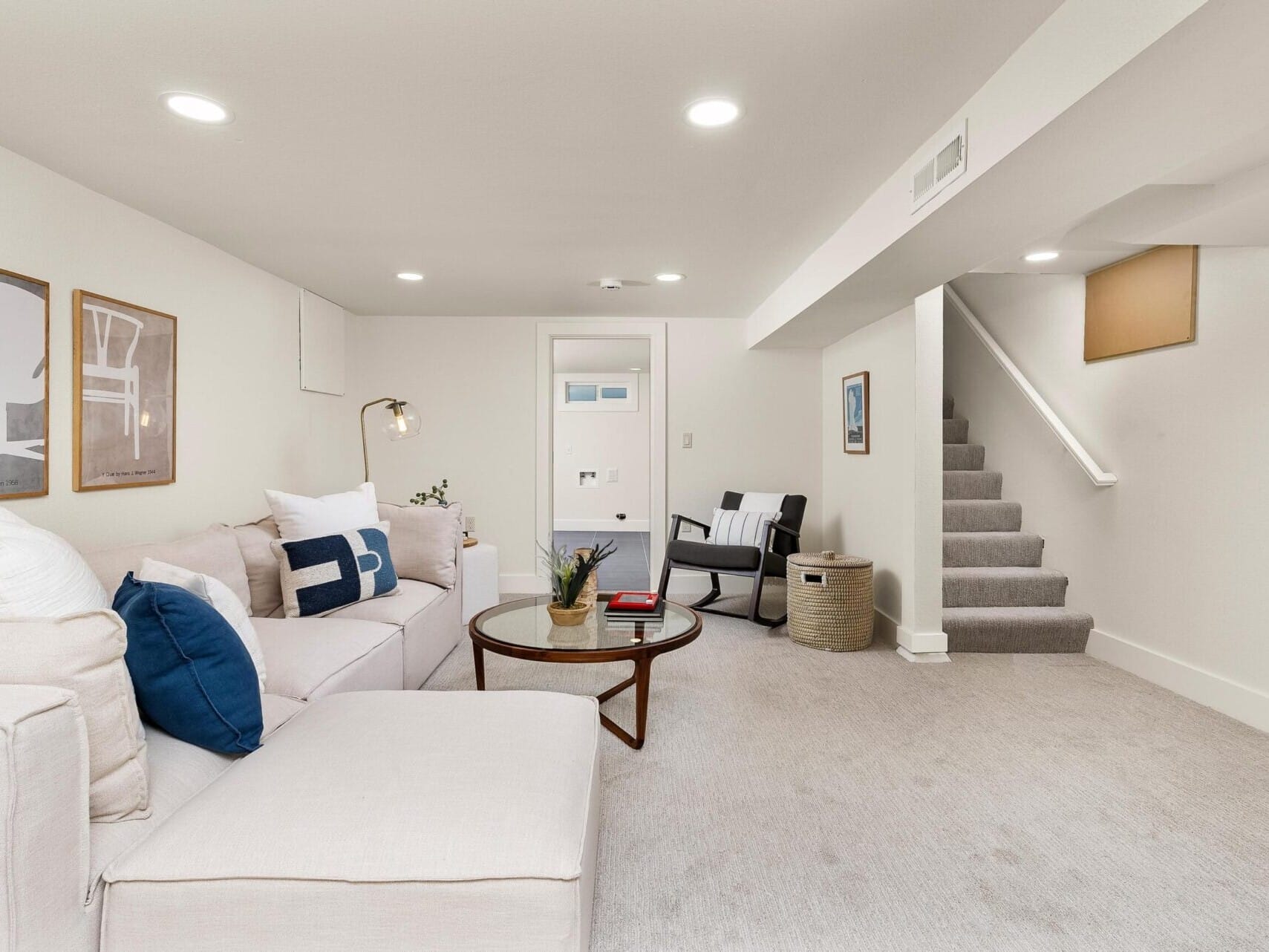 A modern basement living area with white walls and carpet showcases a large beige sectional sofa adorned with blue and white pillows. A round glass coffee table sits in front, completing the inviting space. In the background, a staircase leads upstairs beside a cozy reading nook perfect for any Portland Realtor to unwind.