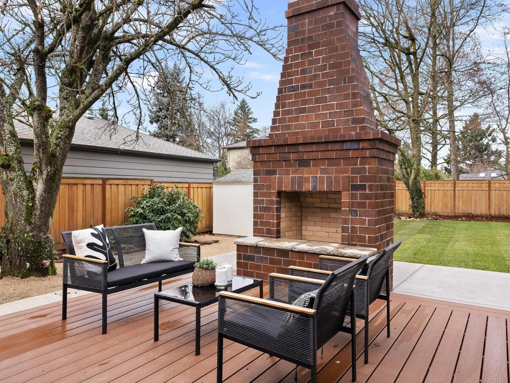 Discover the charm of Portland Oregon real estate with this outdoor patio. It features a wooden deck and a brick fireplace, surrounded by modern black outdoor furniture adorned with white and black cushions. Trees and a wooden fence border under a clear blue sky, offering serene elegance.