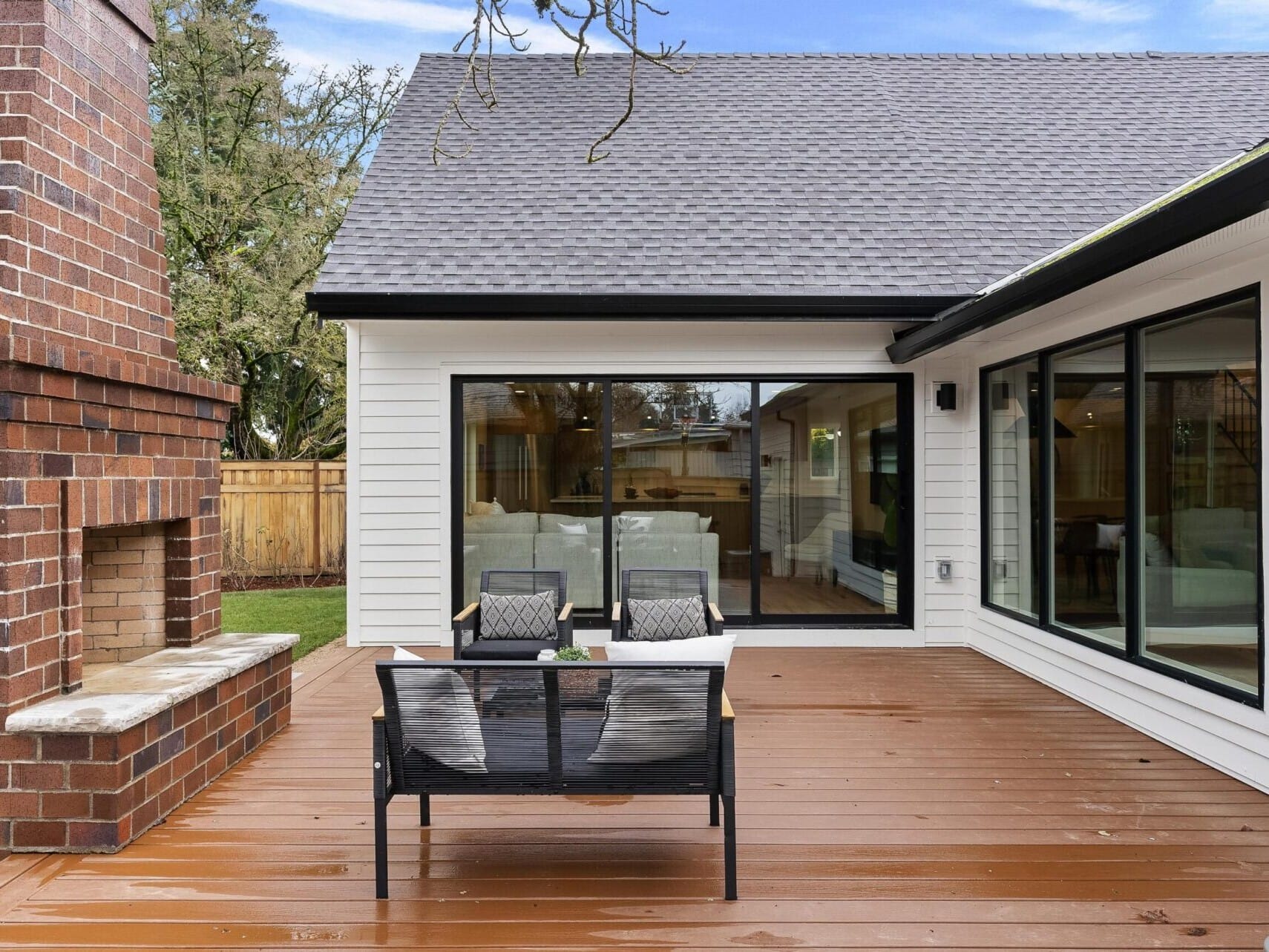 Modern patio with wooden decking featuring a cozy sitting area with two black chairs and a glass table. On the left, there's a large brick fireplace. The house, listed by a top Portland Realtor, boasts white siding and large sliding glass doors that open to the inviting interior.