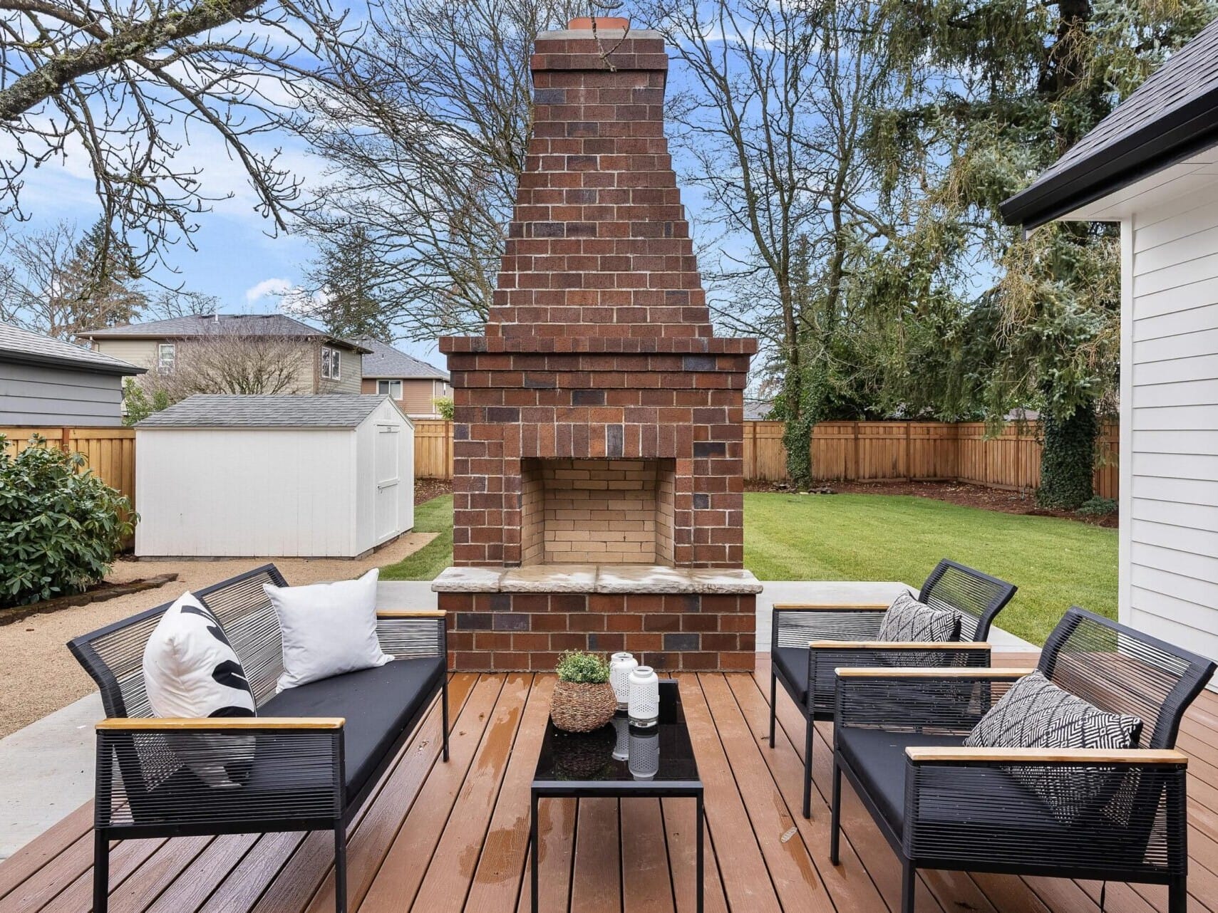 Discover your dream backyard oasis with this outdoor patio featuring a brick fireplace, cozy black seating benches with cushions, and a sleek glass coffee table. Enveloped by trees and a wooden fence, the setting includes a white shed and green lawn. Connect with your trusted Portland Realtor today.