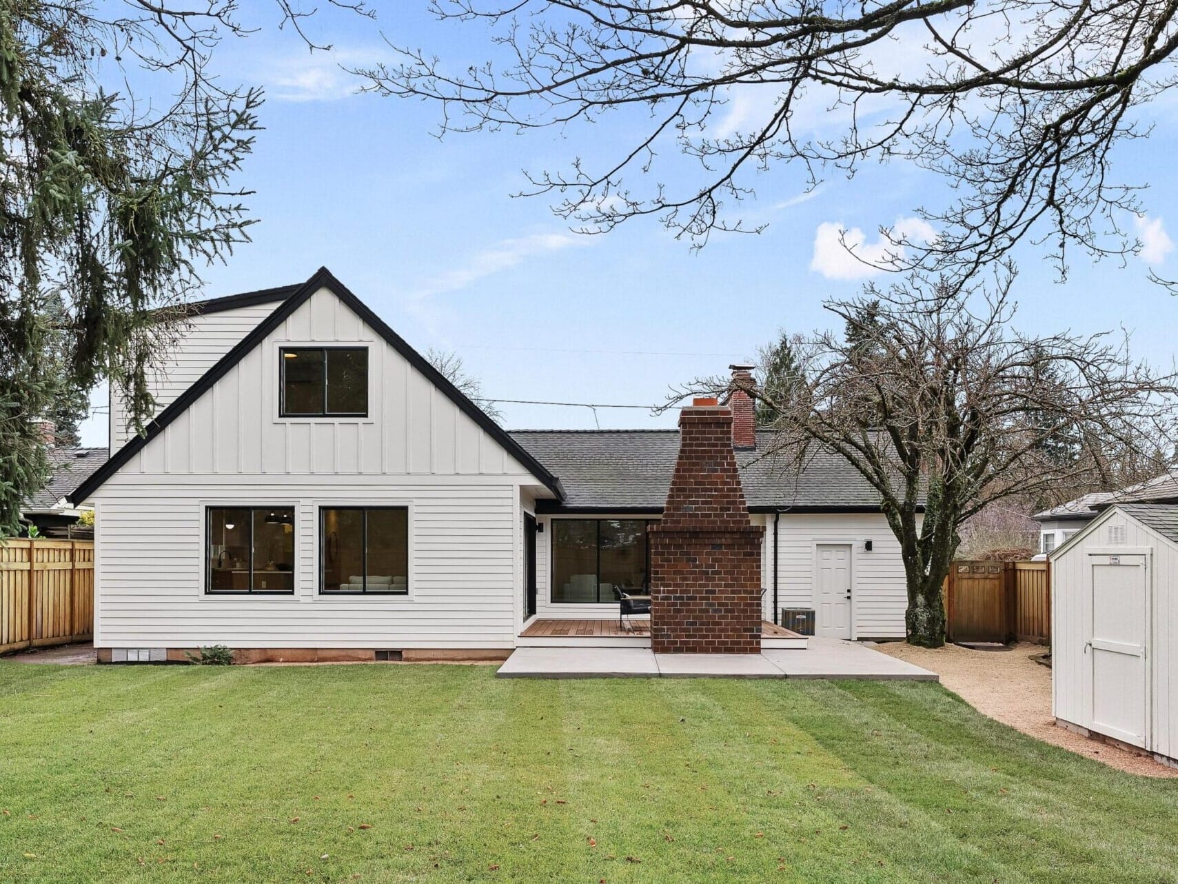 A modern white house with a gabled roof, nestled in Portland's scenic charm, features a spacious backyard and well-maintained lawn. It boasts a large brick chimney, patio seating, surrounded by trees and a wooden fence. A small white shed completes this gem in Portland Oregon Real Estate.