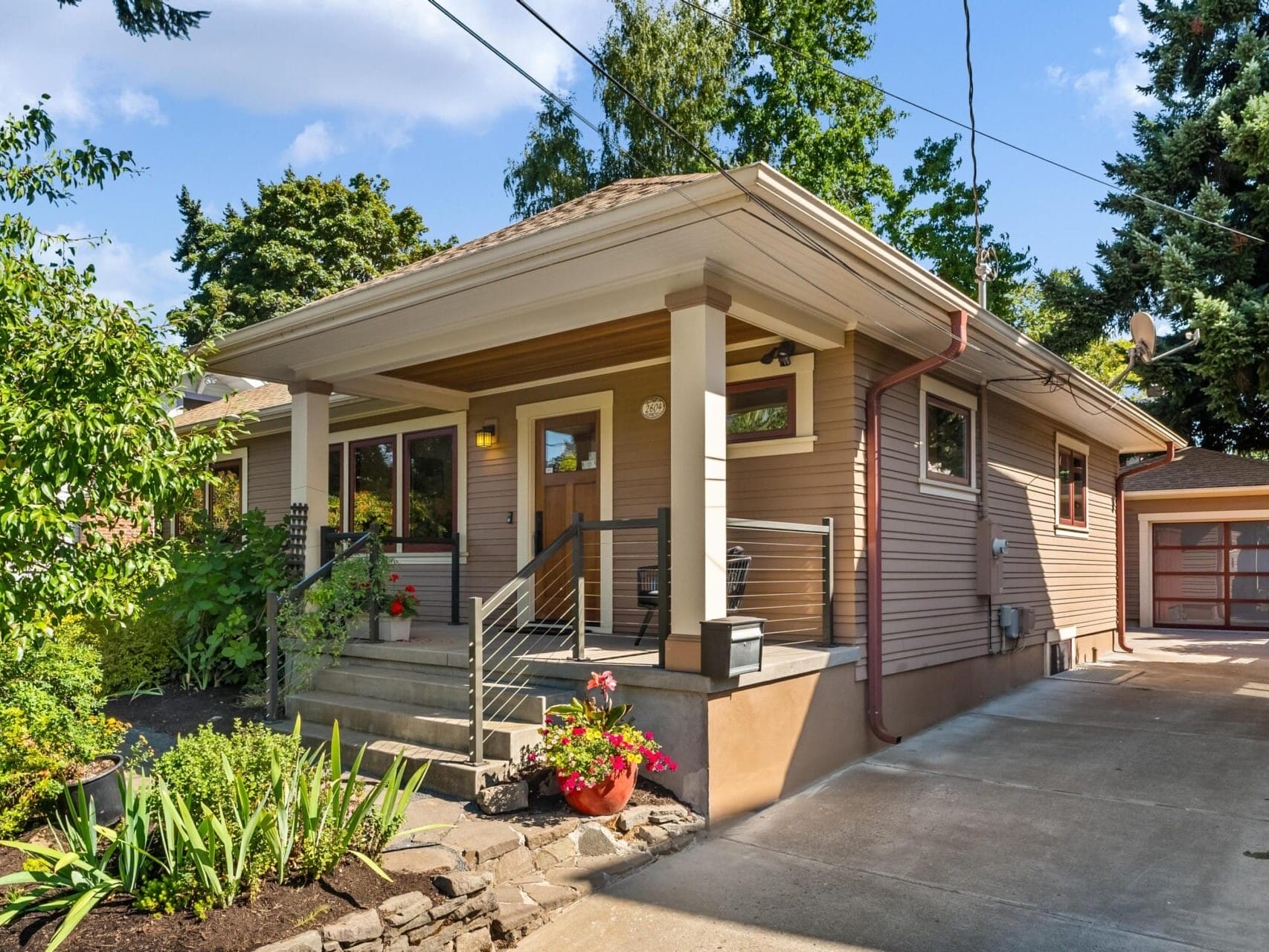 A single-story house in Portland, Oregon, with a covered porch surrounded by greenery. It features beige siding, a paved driveway, and a porch adorned with potted plants. The yard boasts diverse landscaping and a small stone wall under the clear blue sky.