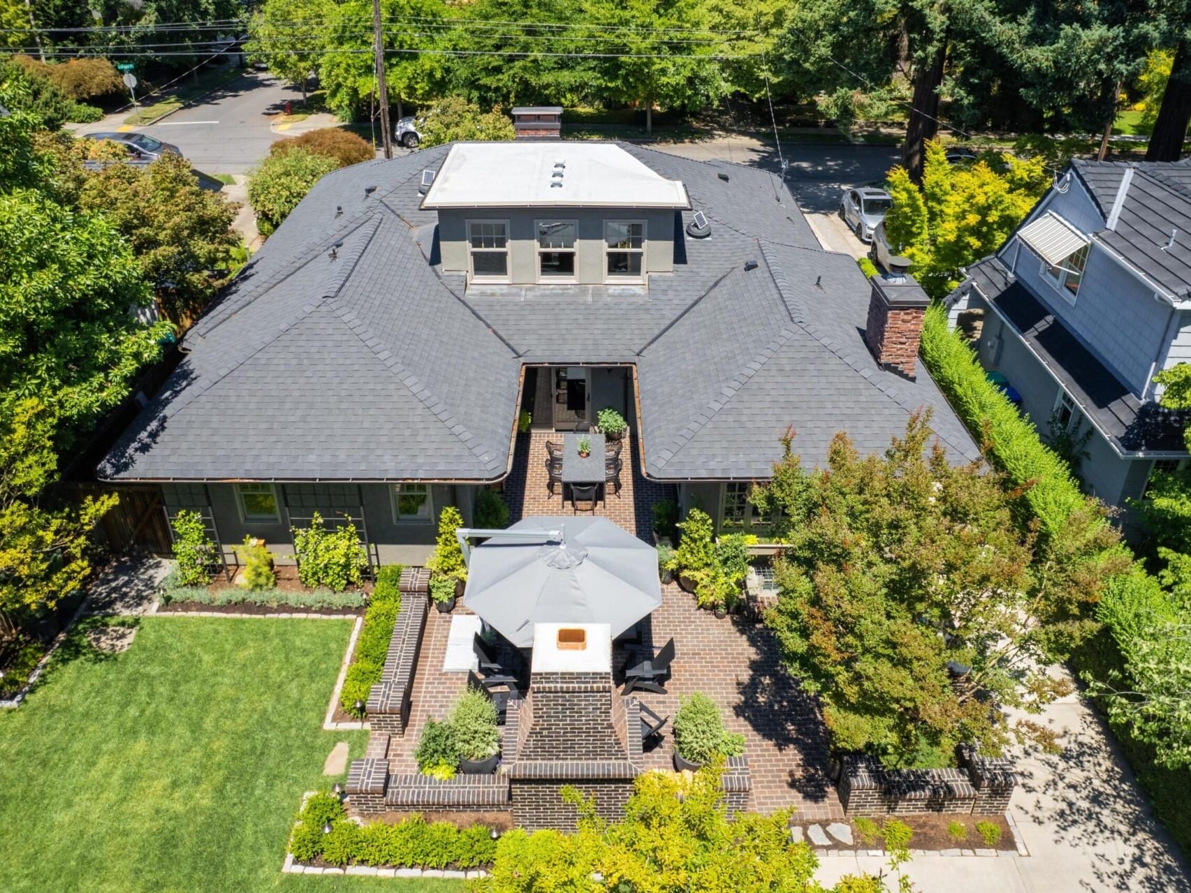 Aerial view of a suburban house in Portland, Oregon Real Estate, boasting a dark shingle roof surrounded by lush greenery. A landscaped garden graces the front with a brick patio area featuring outdoor furniture. Nearby are similar houses and a street nestled in this vibrant community.