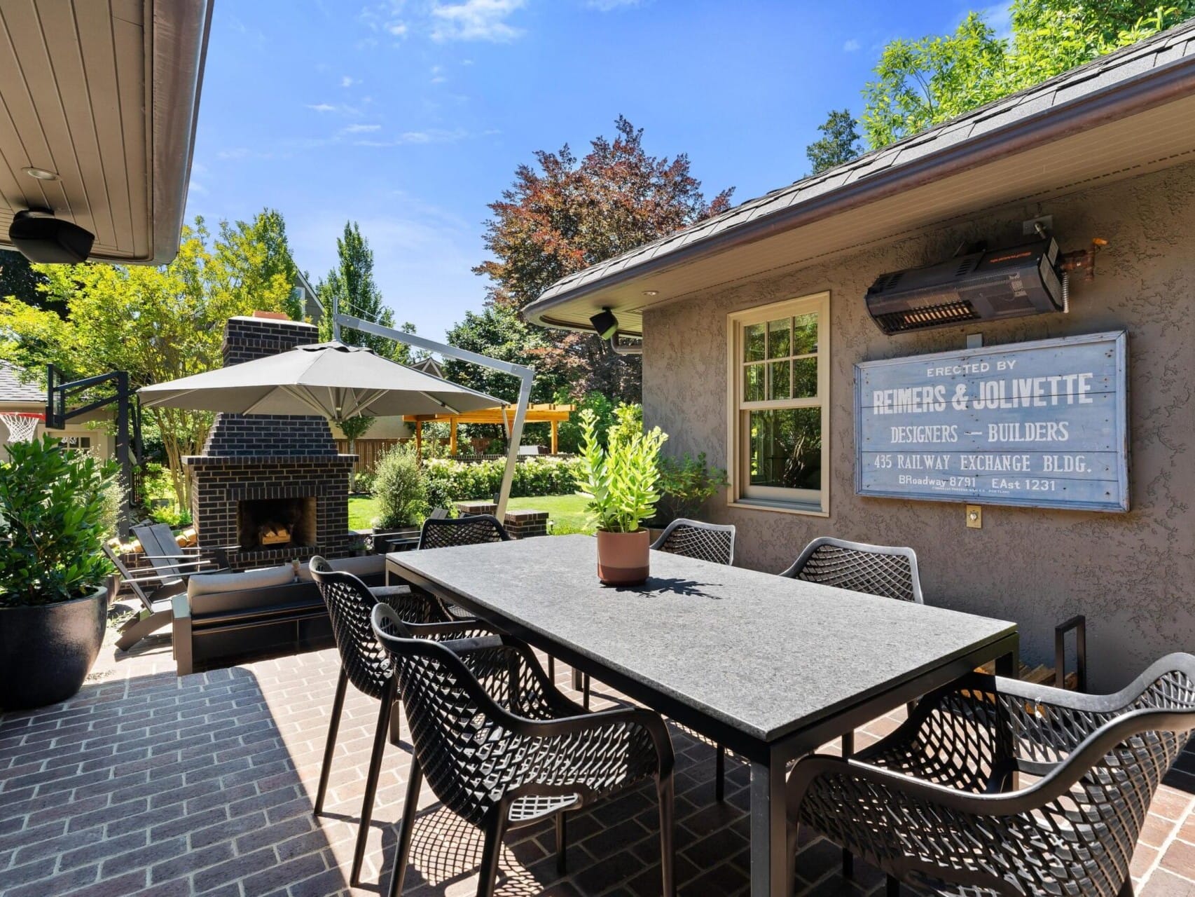 A cozy outdoor patio, perfect for any Portland Oregon real estate gem, features a large dining table with decorative chairs, a brick fireplace, and a generous umbrella. Surrounded by lush greenery for tranquility, it boasts a wall-mounted metal sign that adds rustic charm to the setting.