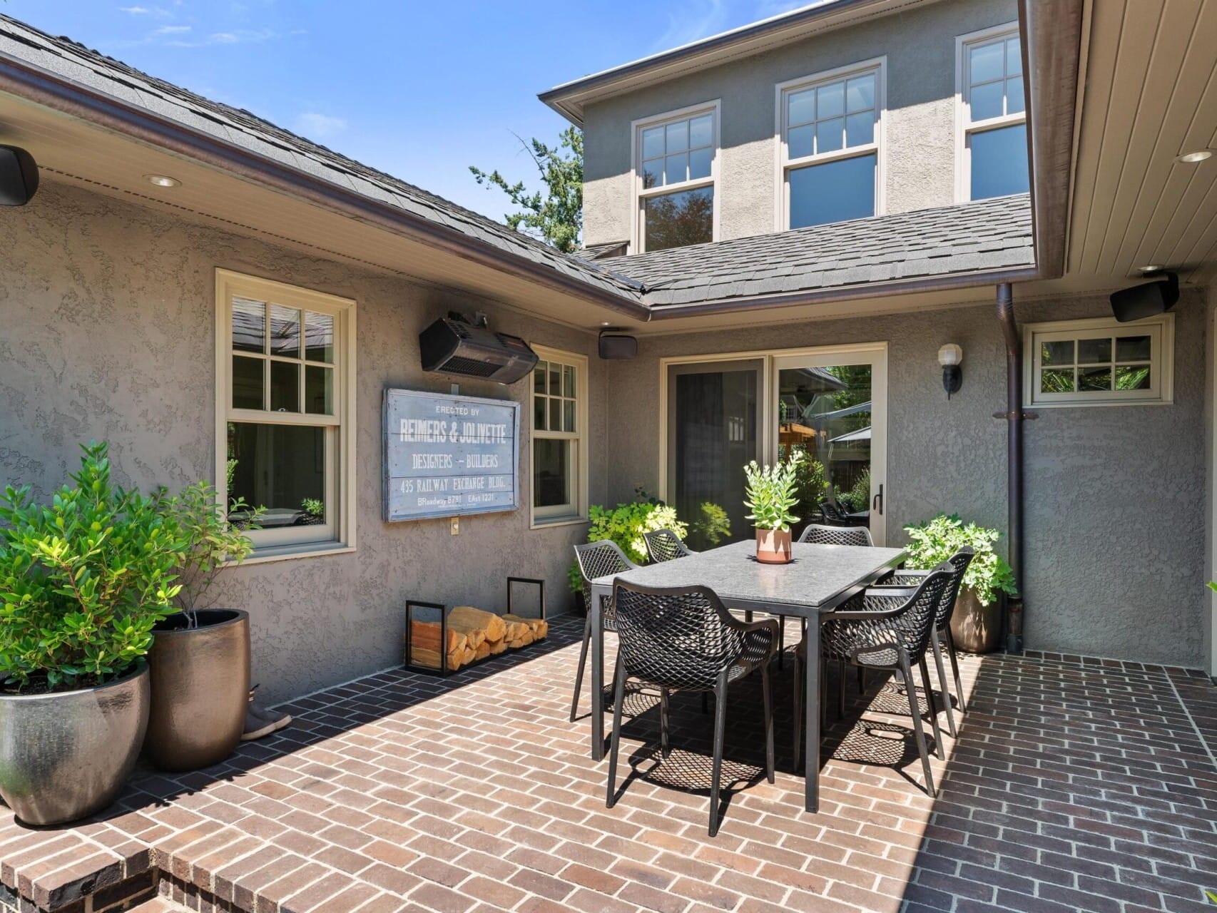 A cozy outdoor patio with a brick floor features a black table and six chairs. Large potted plants and a wooden sign adorn the space, embraced by a gray stone house under a blue sky—a perfect setting that any top Portland Realtor would highlight in sought-after Portland Oregon real estate listings.