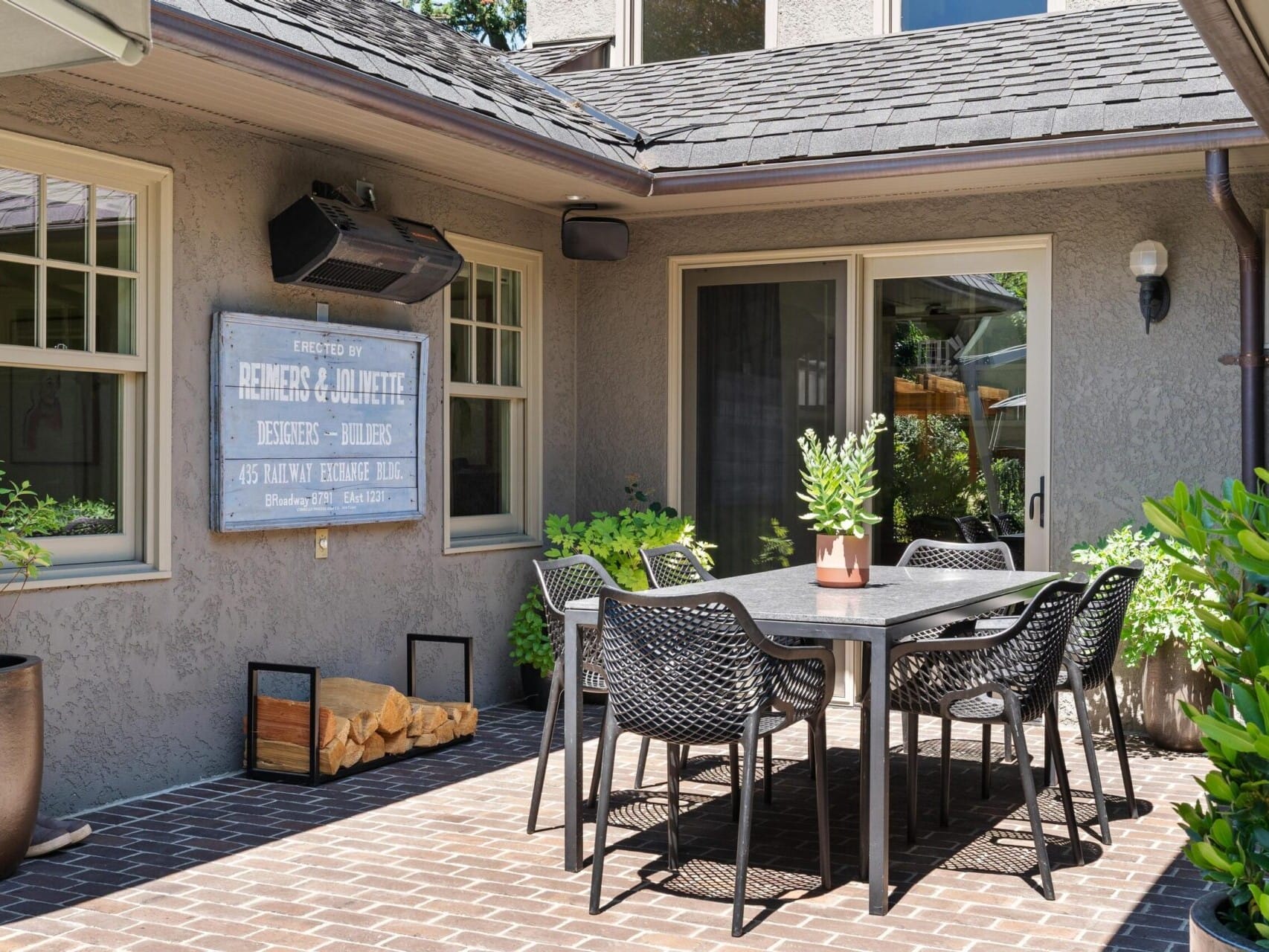 Outdoor patio with a table and six chairs on a brick floor. A large sign is mounted on the wall, surrounded by lush greenery. A glass door leads inside, with a speaker above the sign. This cozy setting exemplifies Portland's charm, attracting interest from those seeking top Portland Oregon real estate.