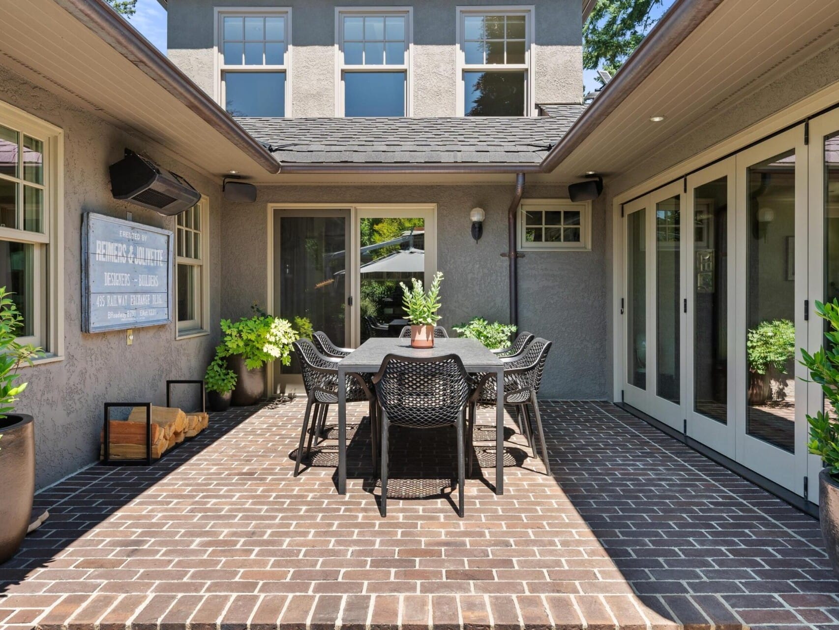 A cozy outdoor patio with a dark wooden dining table and six wicker chairs is nestled in a charming Portland, Oregon real estate gem. Surrounded by potted plants and brick flooring, large windows and a door create a seamless connection between indoor and outdoor areas.