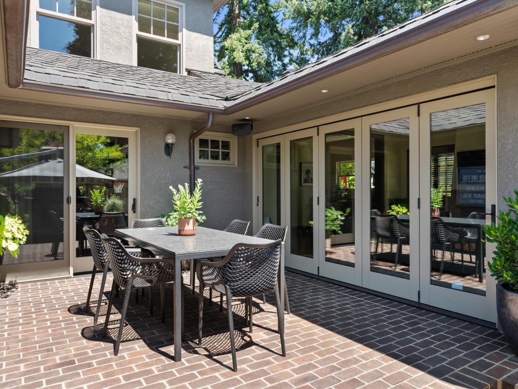 A cozy outdoor patio with a brick floor features a modern dining table and chairs, perfect for enjoying Portland Oregon real estate. Large glass doors lead to the interior, and the area is surrounded by lush greenery. A potted plant decorates the table as bright sunlight filters through the scene.