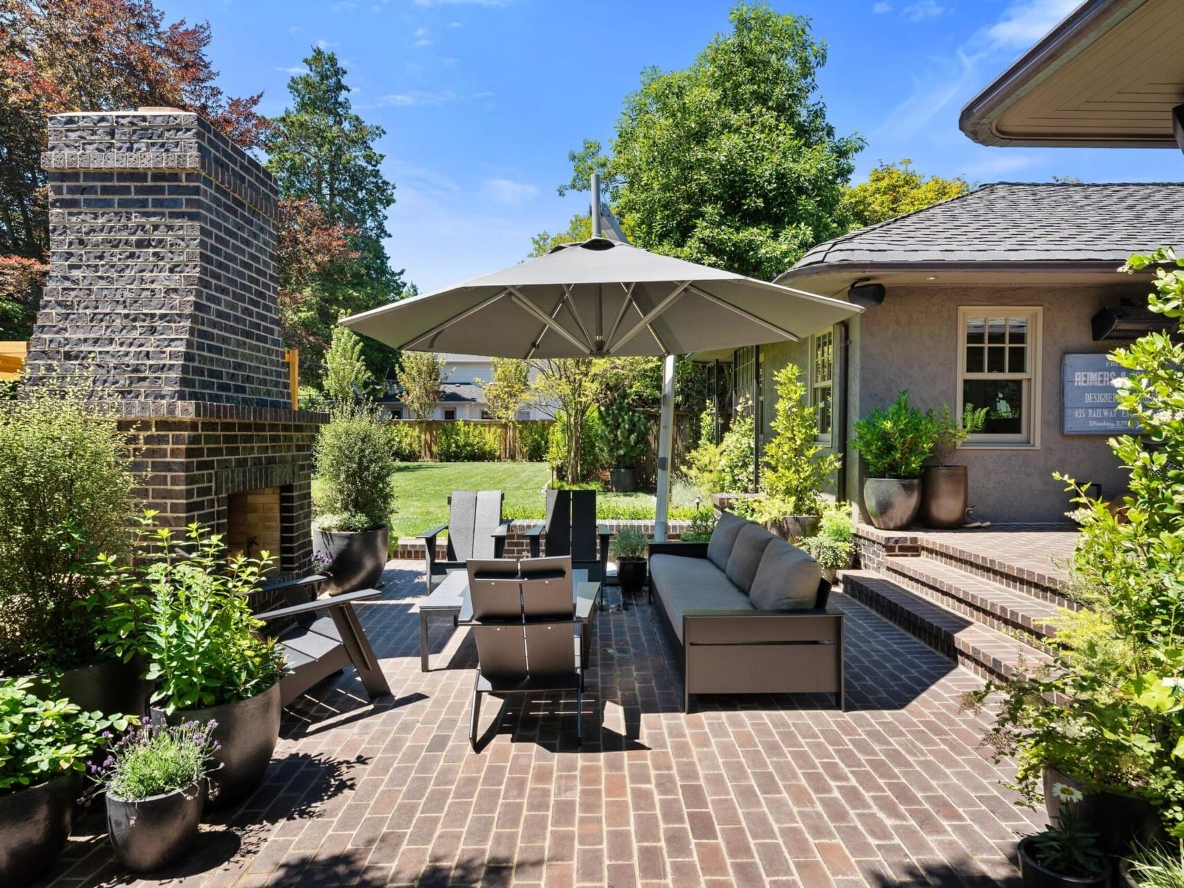 A cozy patio with brick flooring designed by a top real estate agent features outdoor furniture, including a sofa, chairs, and a table under a large umbrella. A tall brick fireplace stands nearby, surrounded by lush greenery and potted plants. The sky is clear, with trees in the background.