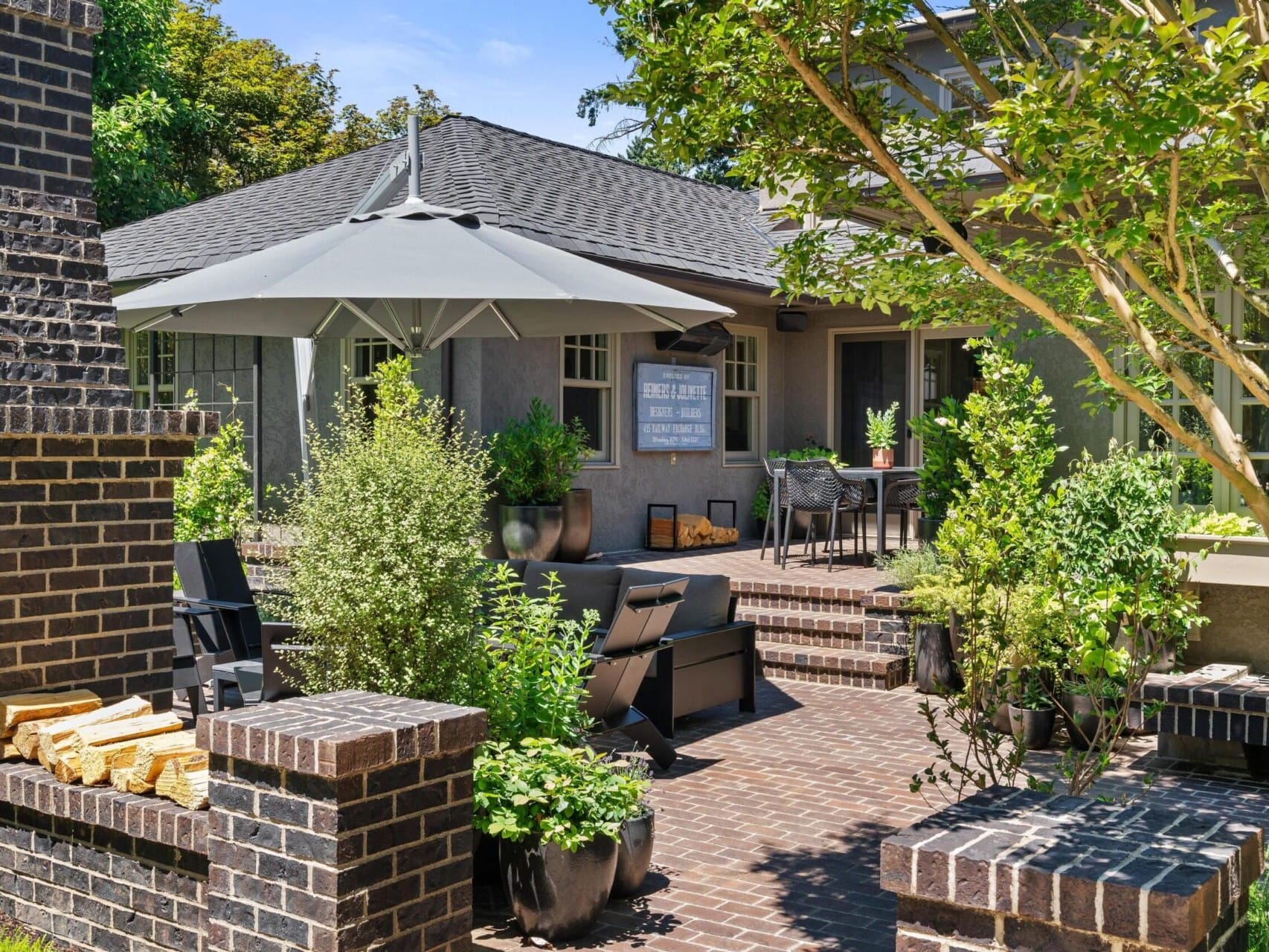 Discover a cozy backyard patio in Portland, Oregon real estate, featuring brick flooring and walls. With a large gray umbrella, outdoor seating, a dining table, and lush green plants, this tranquil space is perfect for relaxation. A small stack of firewood nestled between brick pillars completes the charm.