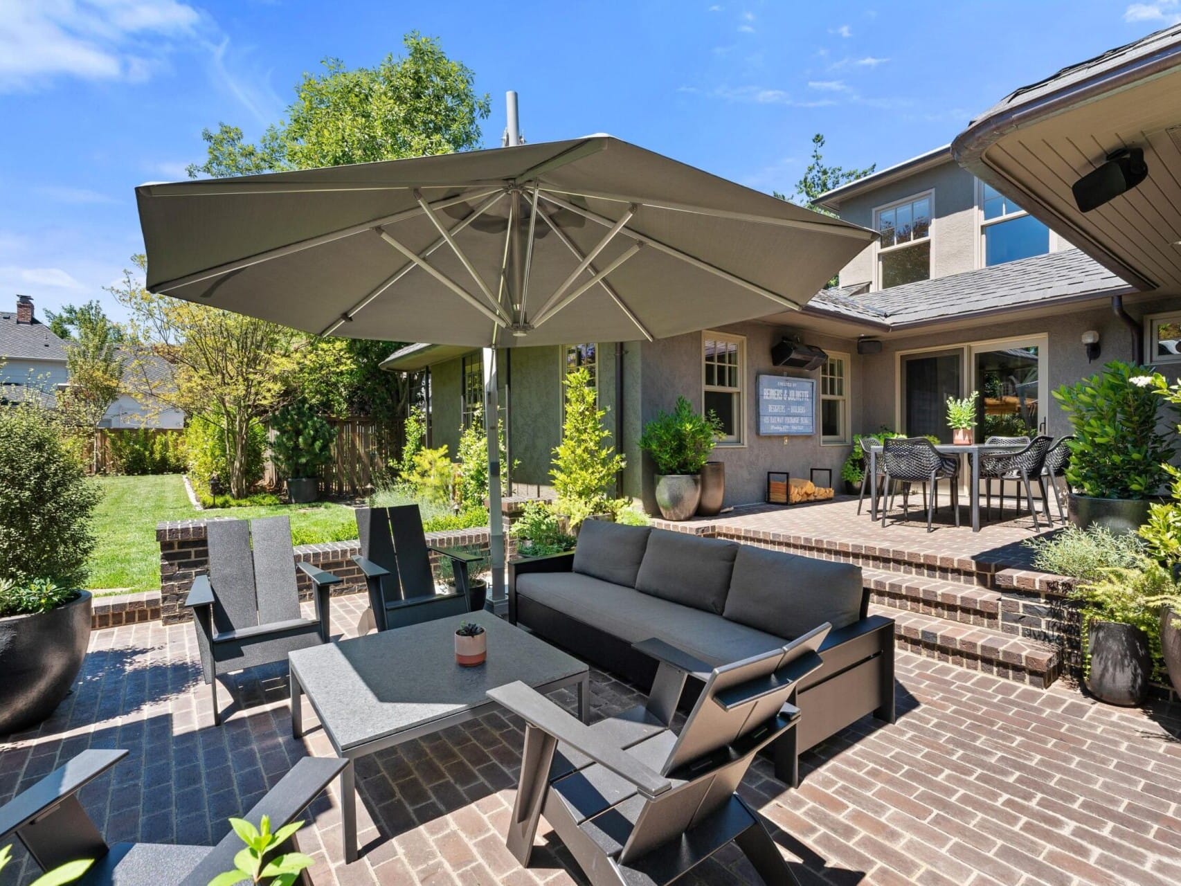 A spacious backyard with a large umbrella, outdoor seating, and a table on a brick patio. This Portland Oregon real estate gem features patio doors and windows surrounded by lush greenery and a manicured lawn under a clear blue sky.