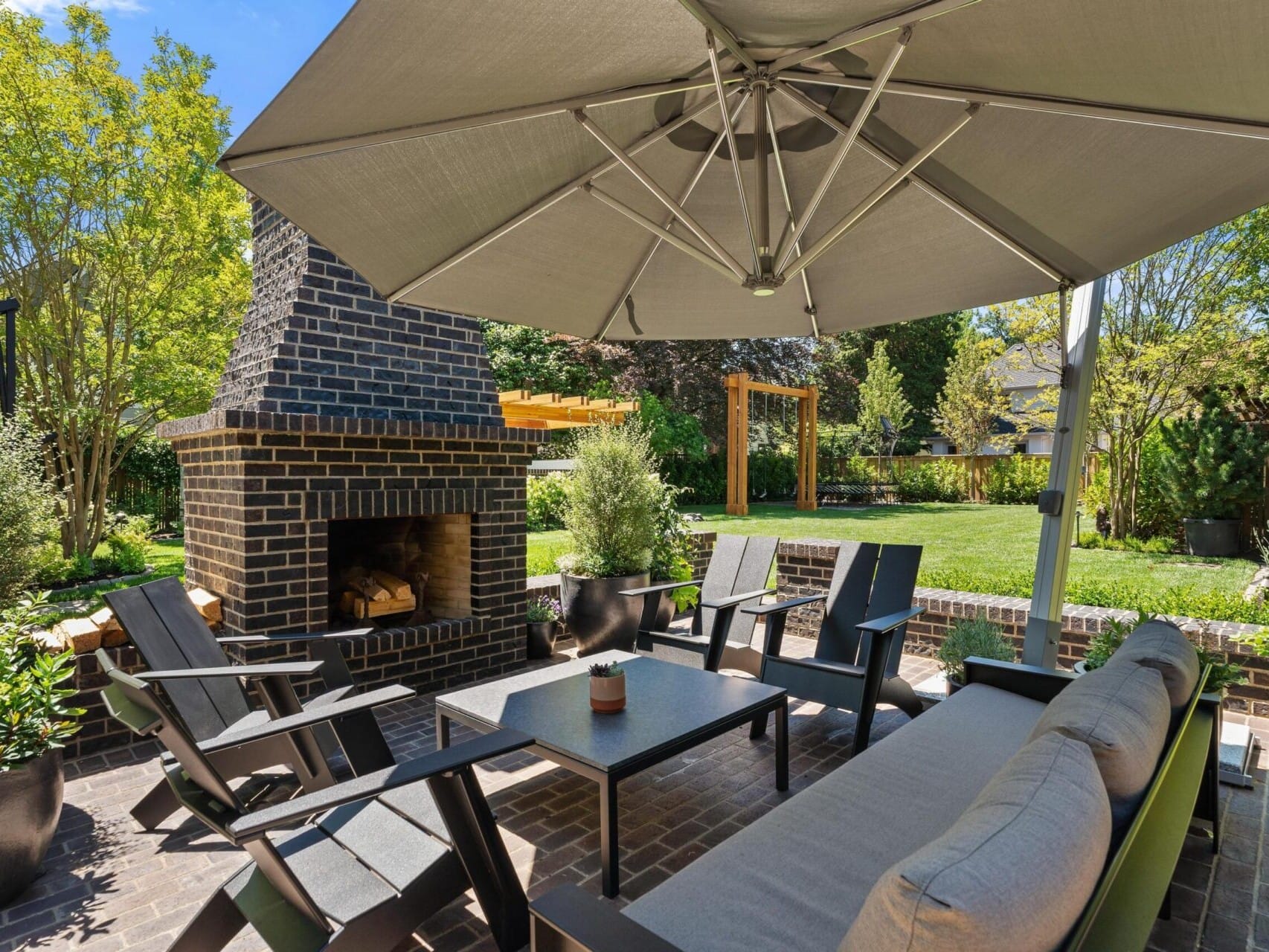 A cozy outdoor patio with a large umbrella shading a seating area offers Pacific Northwest charm. Dark lounge chairs and a sofa encircle the coffee table, while a brick fireplace stands nearby, surrounded by lush greenery—a true Portland Oregon real estate gem.