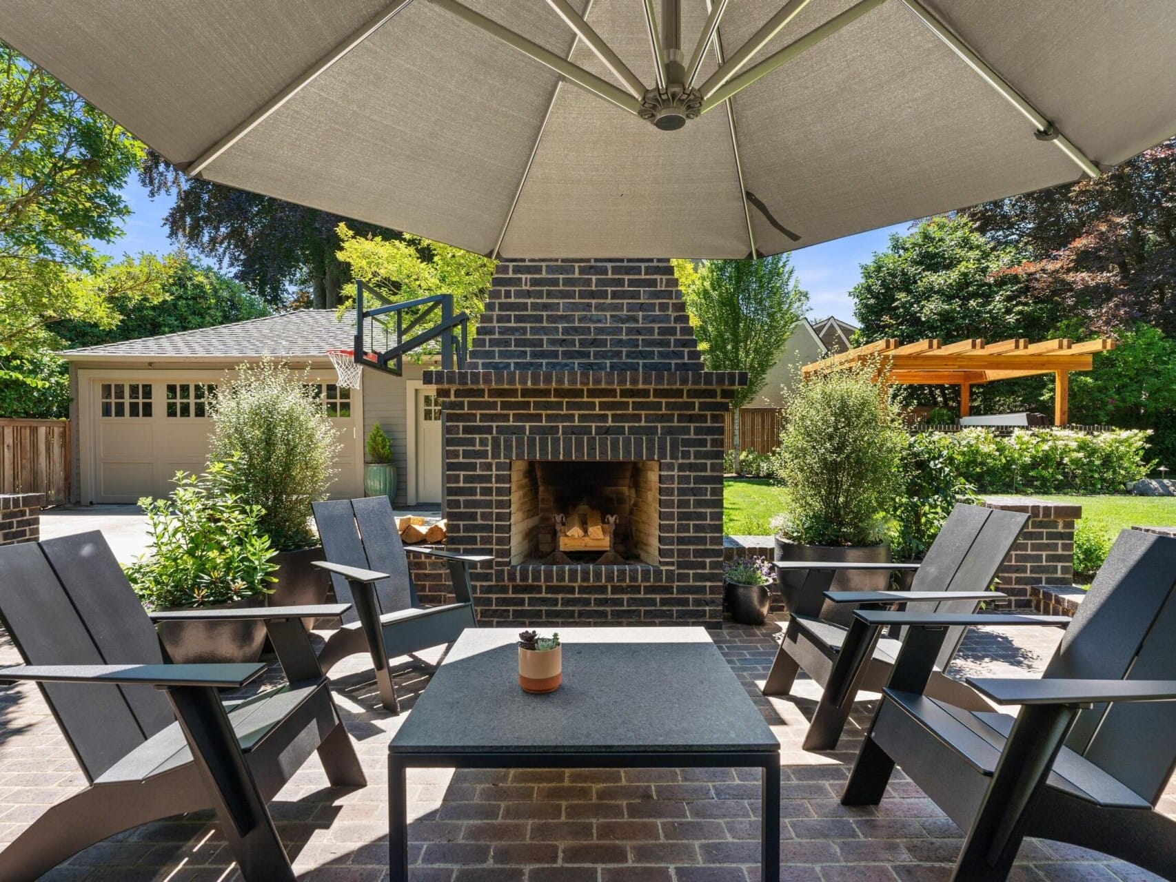 A cozy patio in Portland, Oregon real estate features a brick fireplace surrounded by four black chairs and a central table. A large umbrella provides shade, and potted plants add greenery. In the background, there's a garage and a wooden pergola in a lush garden.