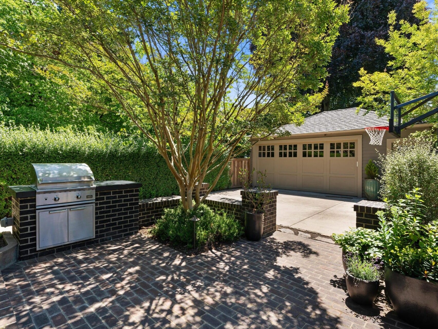 A sunlit backyard in Portland features a brick patio with a built-in stainless steel grill. A large tree provides shade, while a garage with a basketball hoop stands in the background. Lush green plants and hedges surround this oasis, perfect for any Portland Oregon real estate gem.