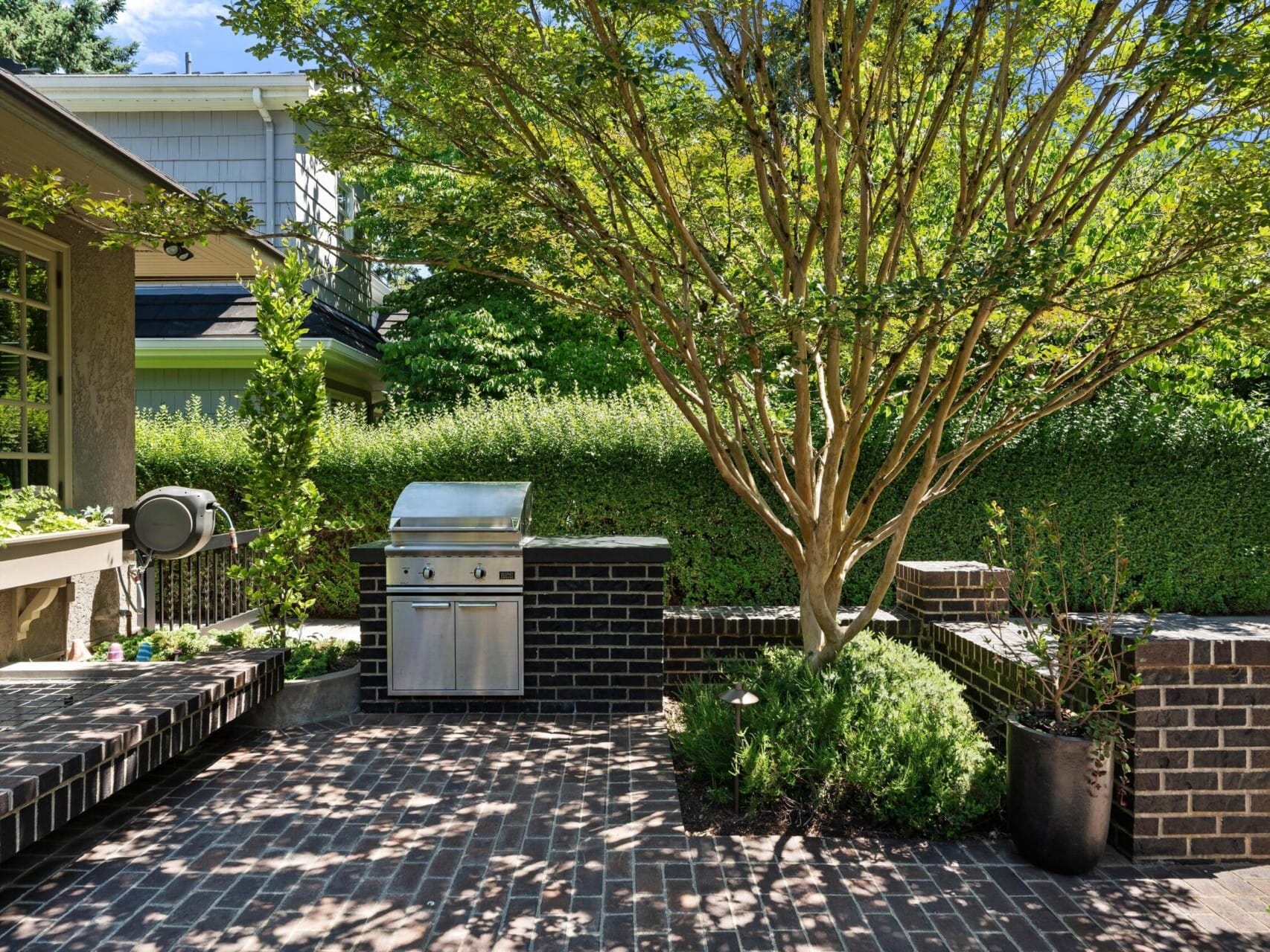 Experience a serene and inviting outdoor patio with a built-in brick grill, where lush greenery and tall hedges offer privacy. A mature tree provides shade over the brick-paved area—a true Portland Oregon real estate gem. Discover more with a top real estate agent today!