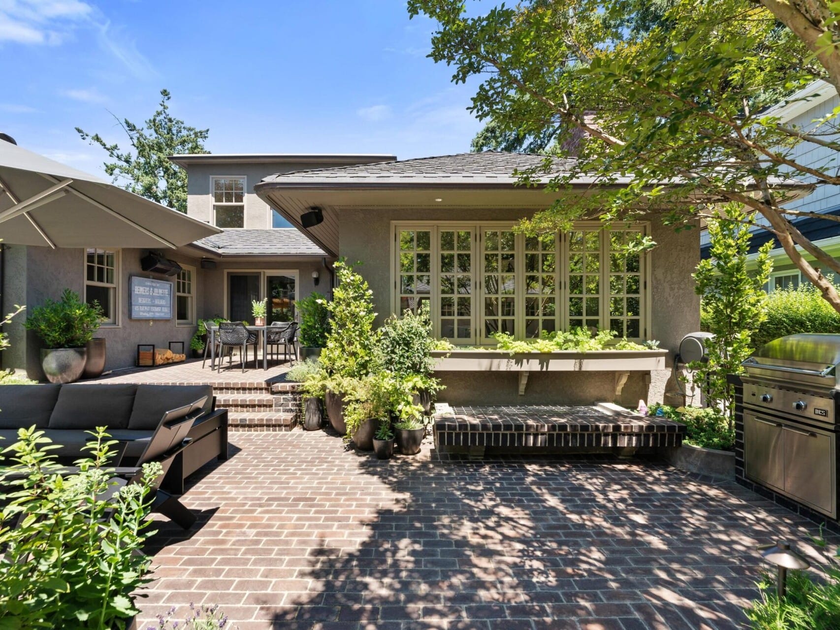 A sunny backyard patio in Portland Oregon real estate showcases a brick floor, a seating area with a dark outdoor sofa, a dining set, and a barbecue grill. Lush greenery surrounds the space, while a large umbrella provides shade. A house with large windows completes this slice of serenity.