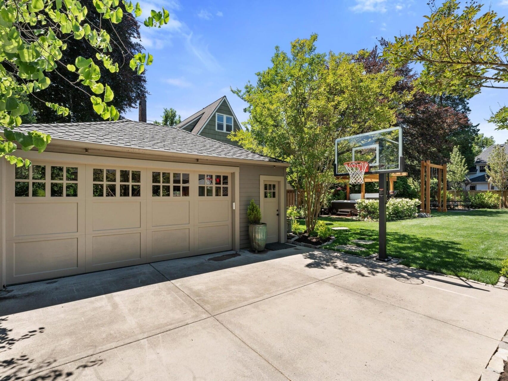 A two-car garage with a light gray door and a basketball hoop graces this Portland, Oregon real estate gem. The landscaped yard boasts trees and shrubs, while a house with a steep roof sits elegantly in the background under the clear blue sky.