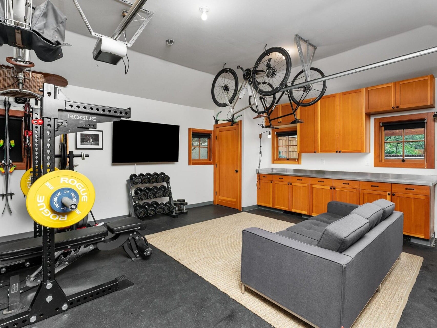 This well-organized home gym features a weightlifting setup, dumbbells, and a bike hanging from the ceiling. Alongside a gray sofa, wall-mounted TV, wooden cabinets, and window views outside, it embodies Portland Oregon Real Estate style and charm.