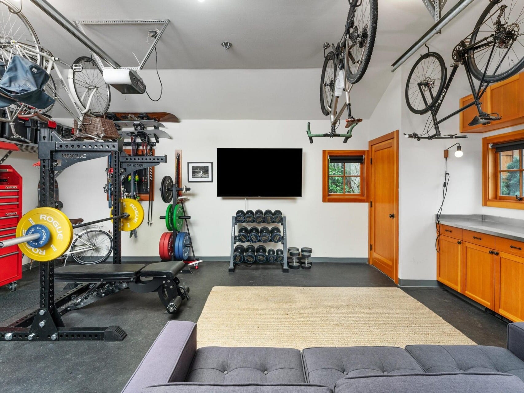 A well-organized home gym in a garage with weightlifting equipment and dumbbells, showcasing Portland Oregon real estate charm. Bicycles hang from the ceiling, while a flat-screen TV is mounted on a white wall beside a gray sofa and wooden cabinets.