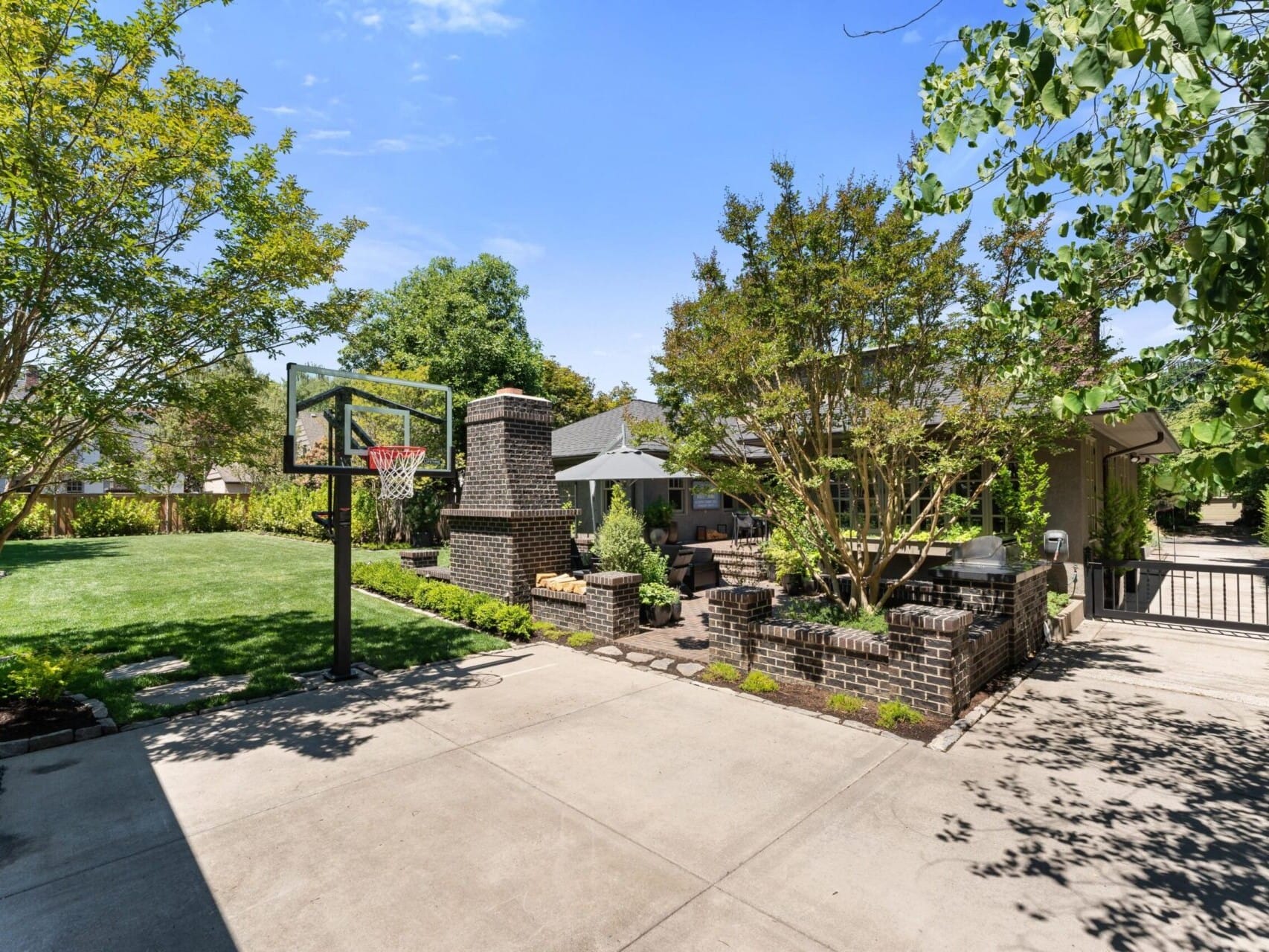 A suburban driveway leads to a house with a lush green lawn and trees, embodying the charm often sought in Portland Oregon real estate. A basketball hoop stands on the left, and a brick outdoor fireplace graces the right. The sunny scene exudes serenity.