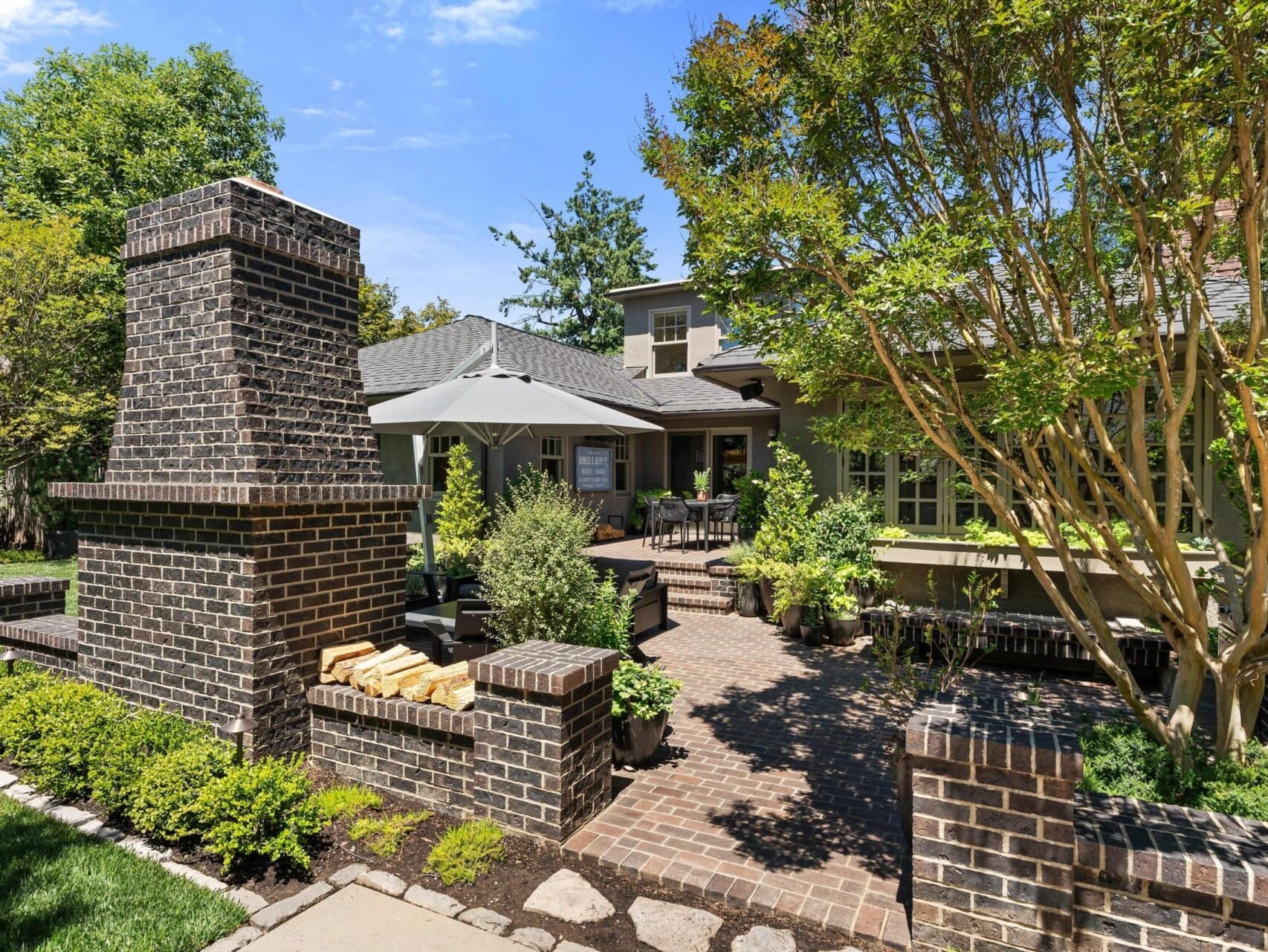 A charming backyard patio in Portland, Oregon real estate features a brick fireplace surrounded by lush greenery and trimmed hedges. The patio includes a seating area beneath a large umbrella, with the house's rustic porch visible in the background—an ideal find for those seeking serenity.