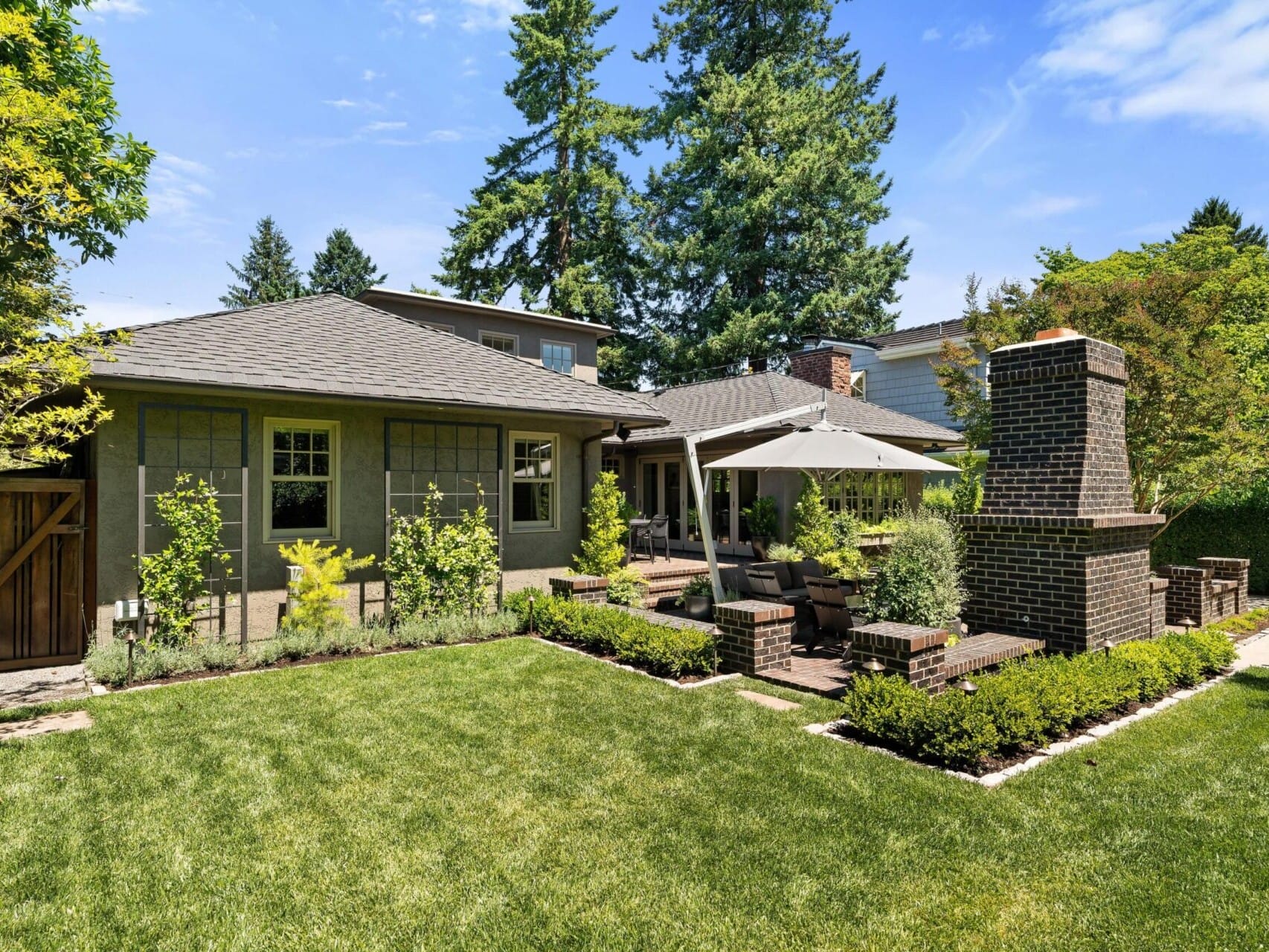 A modern backyard, perfect for Portland Oregon real estate enthusiasts, features a well-manicured lawn, a patio with outdoor seating under a large umbrella, and a brick fireplace. Surrounded by lush trees and greenery, it offers a peaceful and inviting atmosphere.