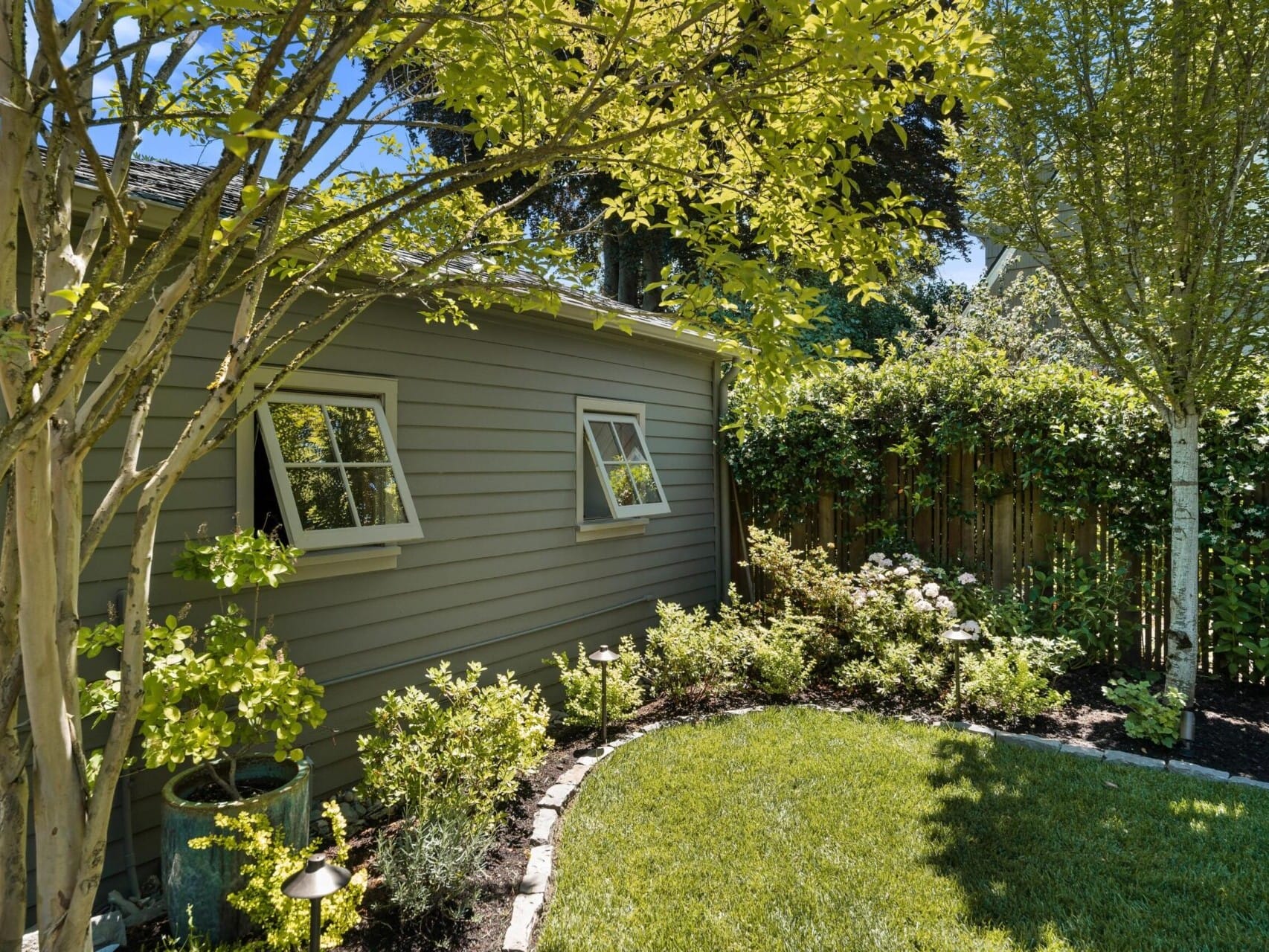 A well-tended backyard, perfect for those seeking Portland Oregon real estate, features a gray shed with two open windows surrounded by lush greenery. A neatly trimmed lawn and a variety of flowering plants create a serene garden setting.