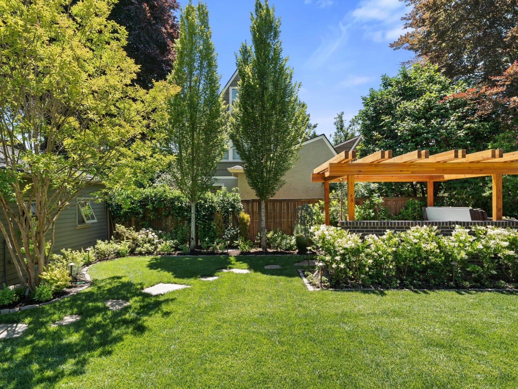 A lush garden with a green lawn, nestled in Portland Oregon real estate, surrounded by tall trees and bushes. A wooden pergola on the right offers a cozy seating area beneath it, set against a backdrop of a light-colored house and clear blue sky.