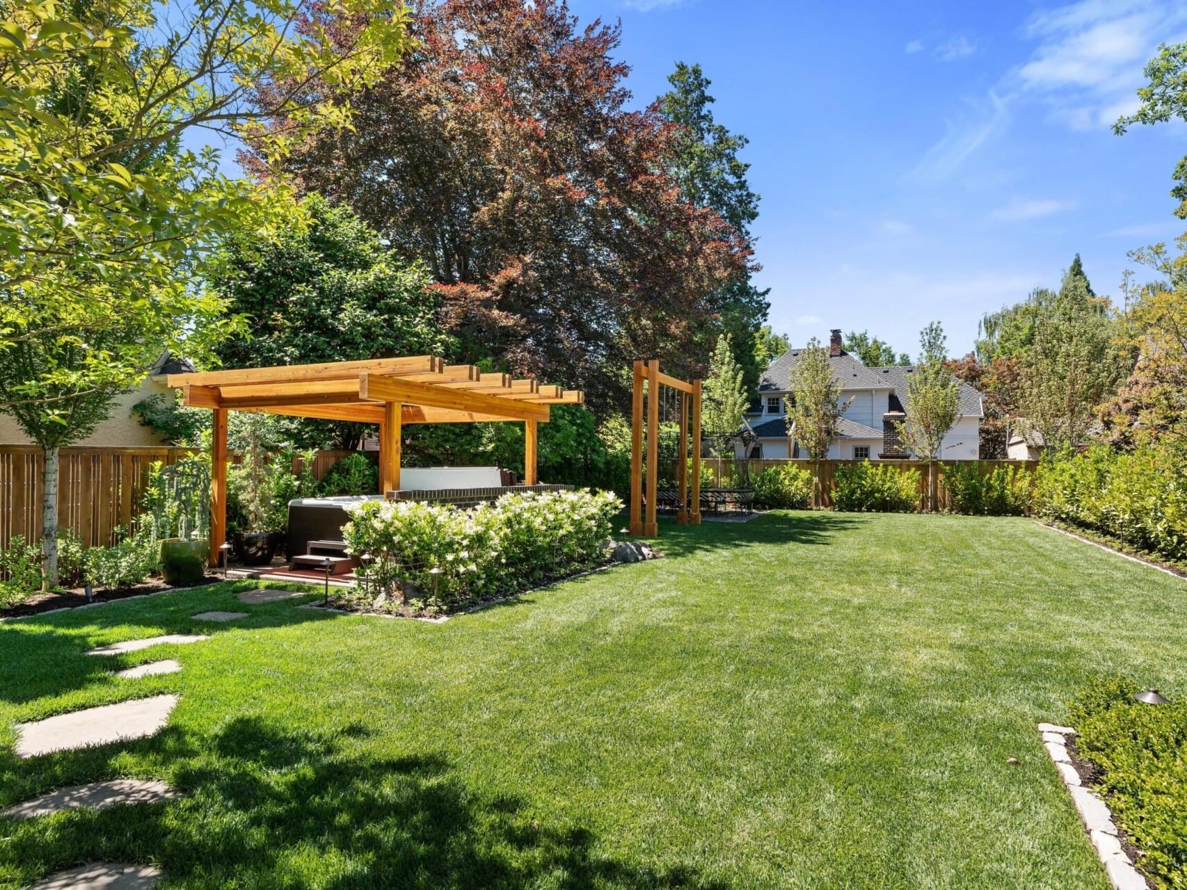 A well-maintained backyard garden with lush green grass and a wooden pergola over a sitting area is surrounded by trees and shrubs. A stepping stone path leads toward the pergola, ideal for Portland Oregon real estate seekers. Houses are visible in the background under a clear blue sky.