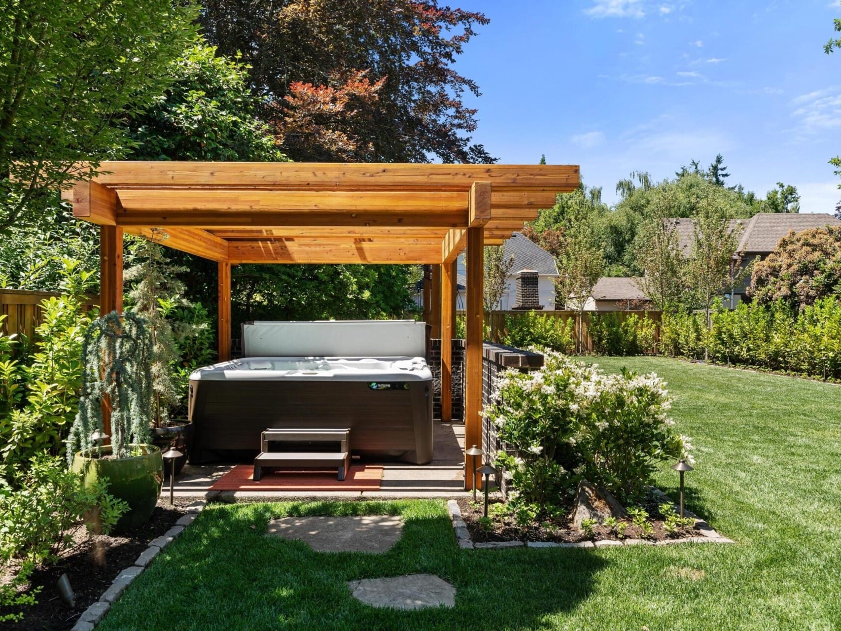 A backyard scene features a wooden pergola covering a hot tub, perfect for relaxing in your Portland Oregon real estate oasis. The surrounding area is landscaped with green grass, shrubs, and trees. Houses and a clear blue sky are visible in the background.