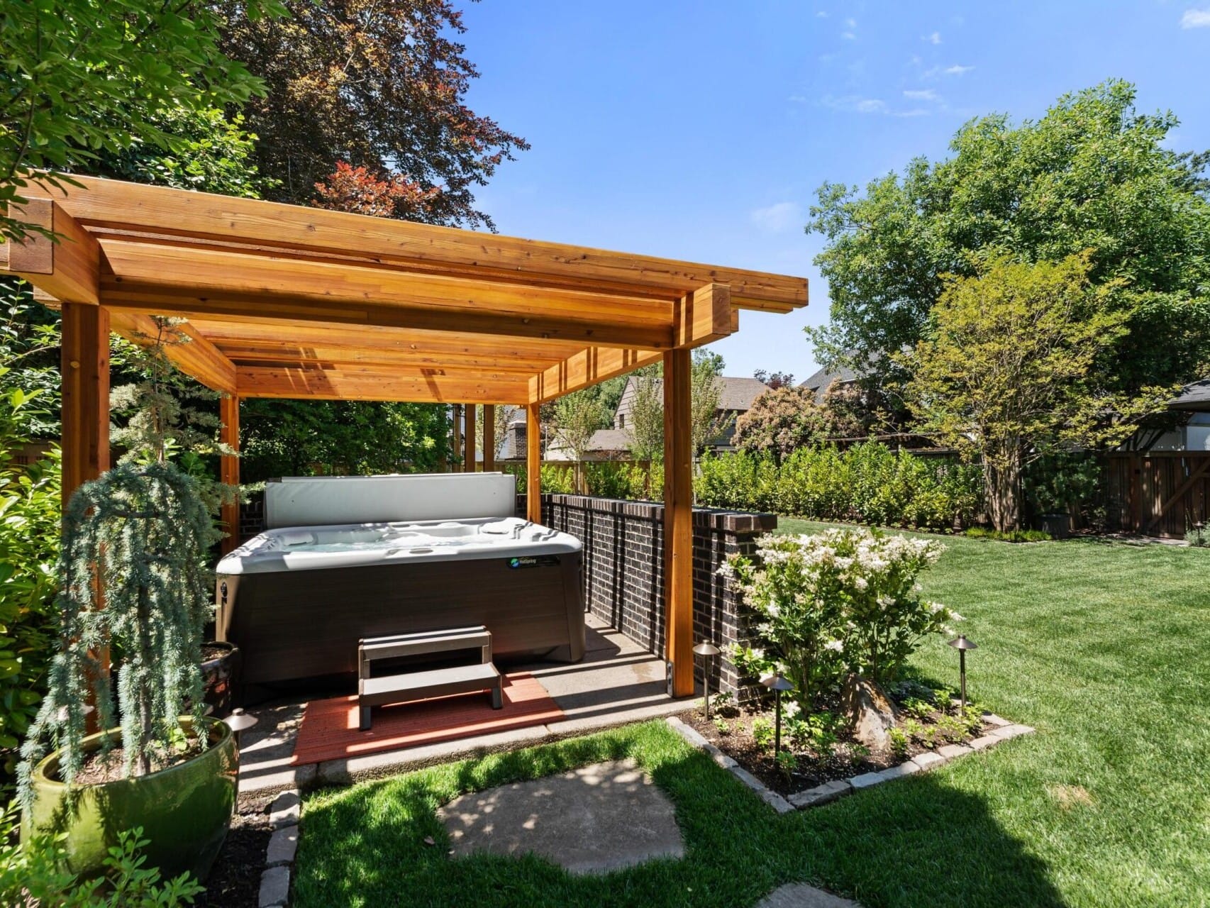 A backyard garden in Portland features a covered wooden pergola sheltering a hot tub. Lush green grass and various plants surround the area, while trees provide natural shade. A pathway leads to the pergola, creating a serene outdoor space—a gem in Portland Oregon's real estate offerings.
