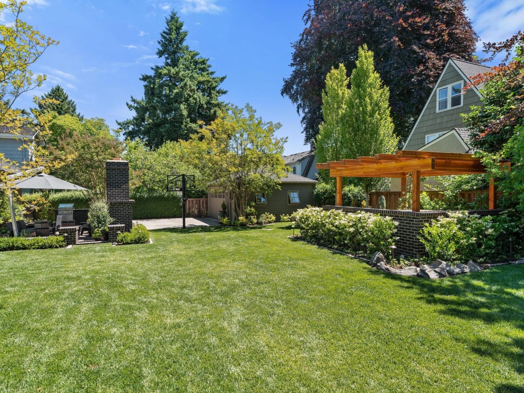 A spacious backyard with a well-manicured lawn is surrounded by trees and shrubs, perfect for enjoying Portland Oregon real estate. There's a wooden pergola on the right, a patio set with an umbrella on the left, and houses in the background under a clear blue sky.