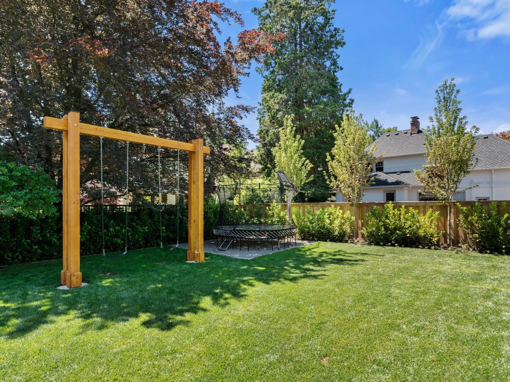 A charming backyard with a wooden swingset, surrounded by a lush green lawn, is perfect for enjoying Portland's beautiful weather. With trees and bushes lining the fence and a trampoline in the corner, this oasis offers an ideal setting in Portland Oregon's real estate market.