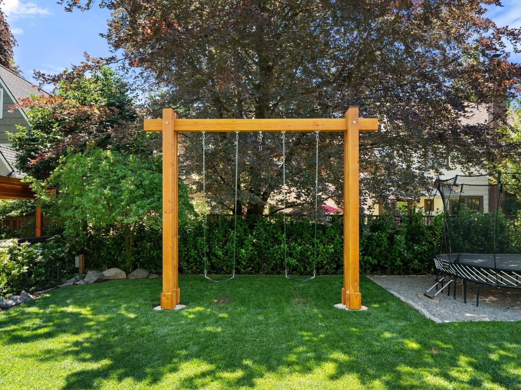 A backyard with a wooden swing set holding two swings, surrounded by green grass and tall trees, offers an idyllic family retreat. A trampoline is visible on the right side, set against a backdrop of charming houses—a perfect slice of Portland Oregon real estate.