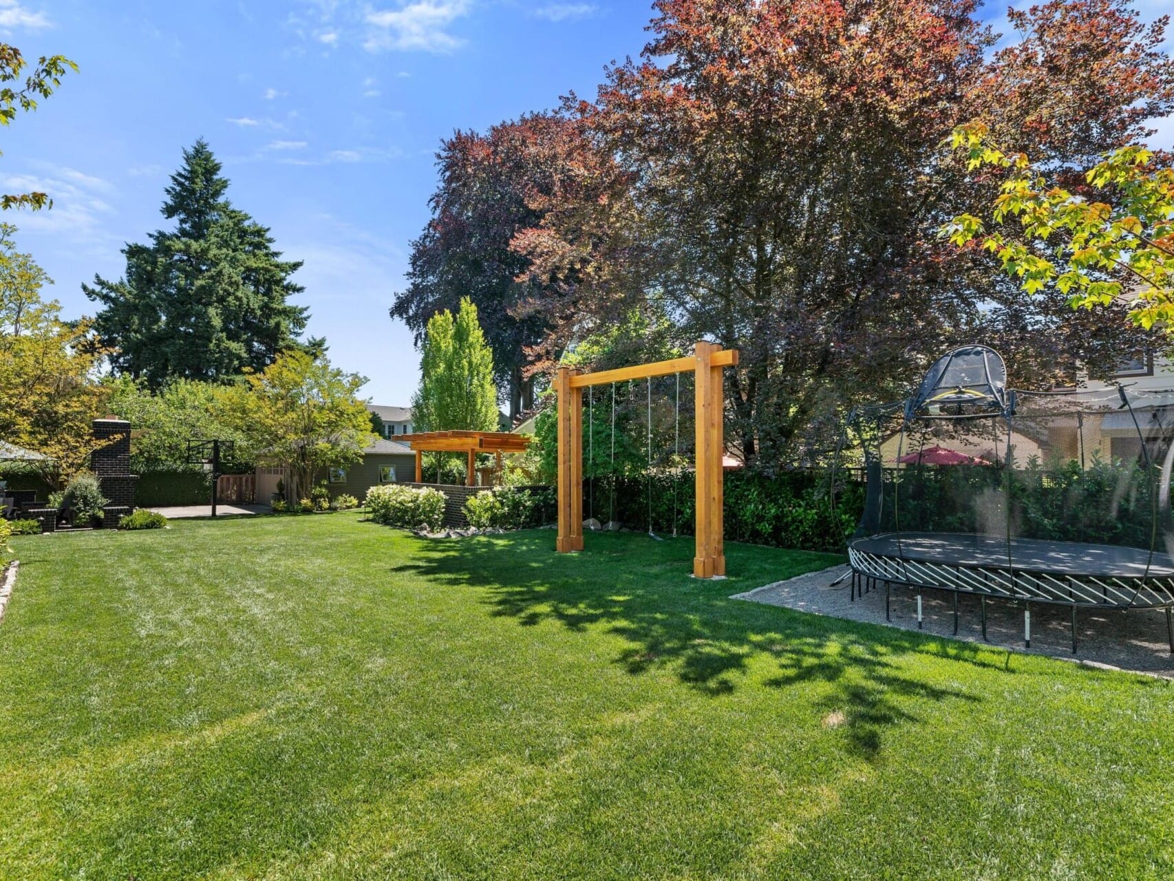 A spacious backyard features a green lawn, a wooden swing set, and a trampoline. Mature trees provide shade, while a small gazebo is visible under Portland's clear blue sky. Perfectly located for those exploring Portland Oregon real estate opportunities with your top real estate agent.