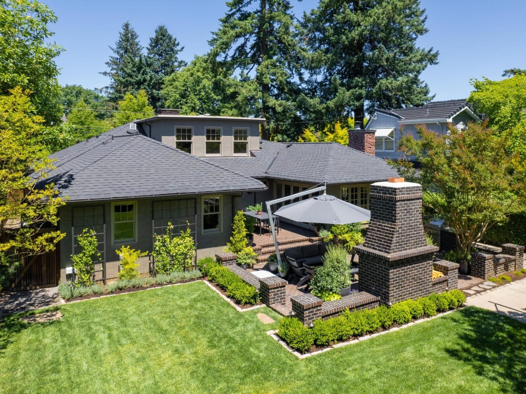 Aerial view of a charming house nestled in Portland, Oregon real estate. The backyard boasts a well-manicured lawn, a brick patio with outdoor seating, and tall trees framing the picturesque scene under a clear blue sky. Ideal for anyone seeking serenity in this vibrant city.