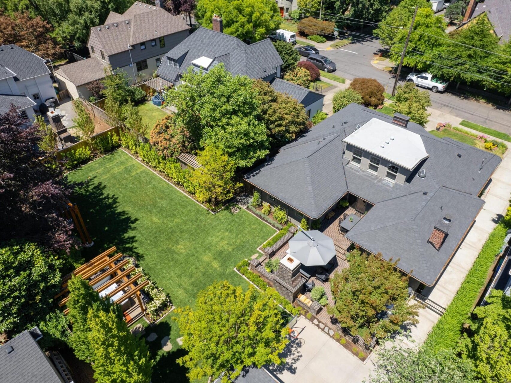 Aerial view of a suburban Portland Oregon real estate neighborhood showcasing a large house with a dark roof nestled amid lush greenery. The property features a well-maintained lawn, garden with pergola, and paved pathway. Nearby houses and tree-lined streets complete this charming scene.