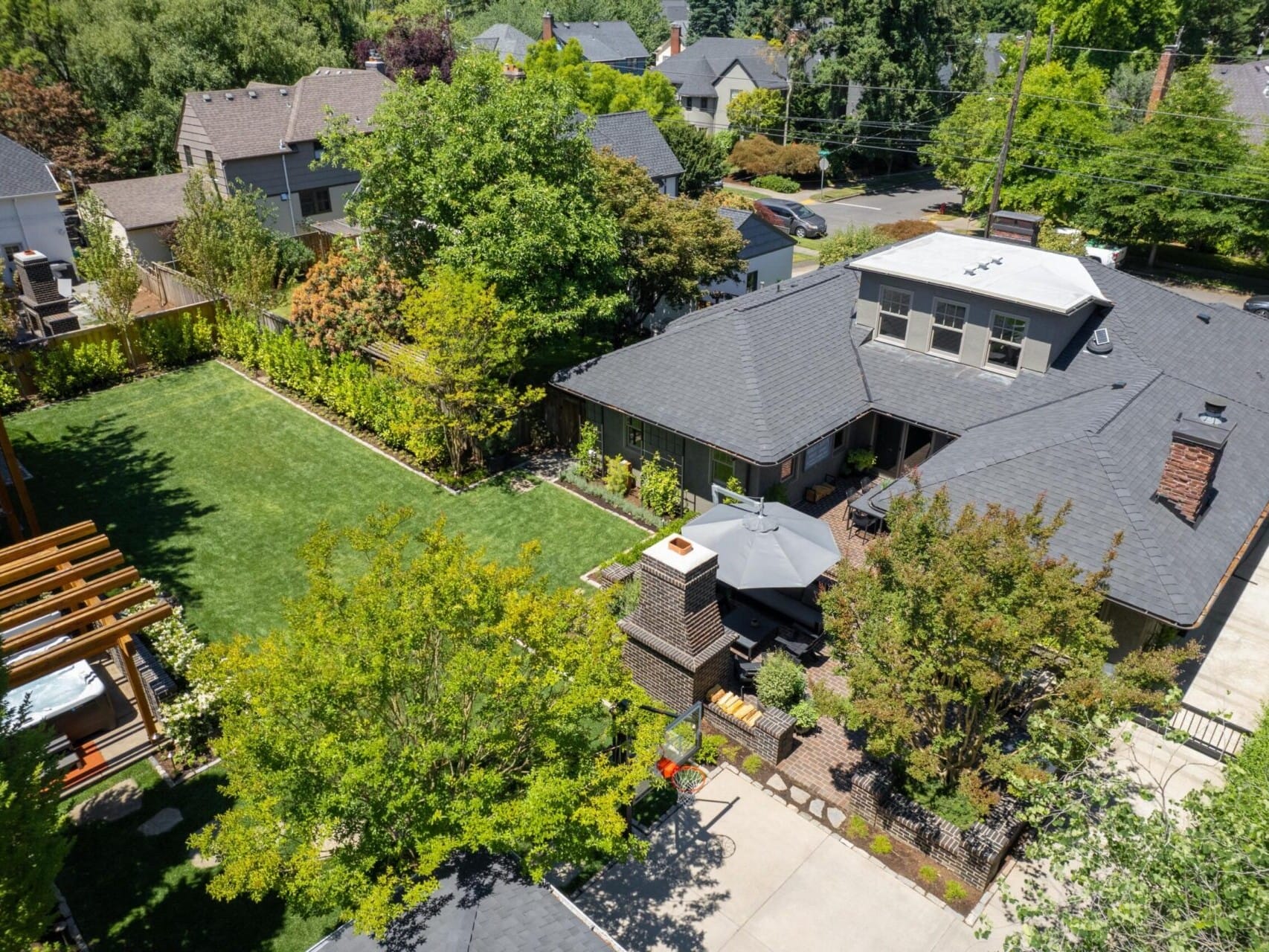 Aerial view of a suburban house with a gray roof, nestled in Portland, Oregon real estate, surrounded by lush green trees and a neatly manicured lawn. A patio area with outdoor furniture and a small garden grace the backyard, while neighboring houses are partially visible.