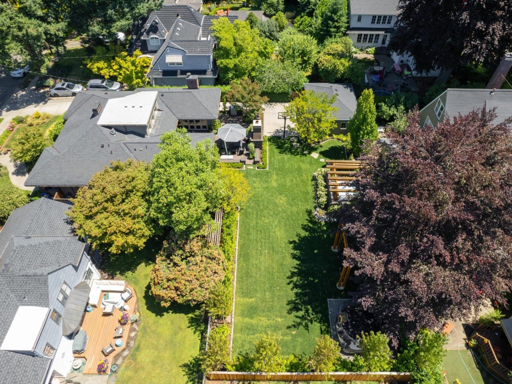 Aerial view of a Portland Oregon real estate gem, showcasing a residential neighborhood with houses boasting grey roofs and a sprawling grassy backyard. Bordered by trees and a wooden fence, the area features patios with outdoor furniture, creating an inviting setting surrounded by lush greenery.