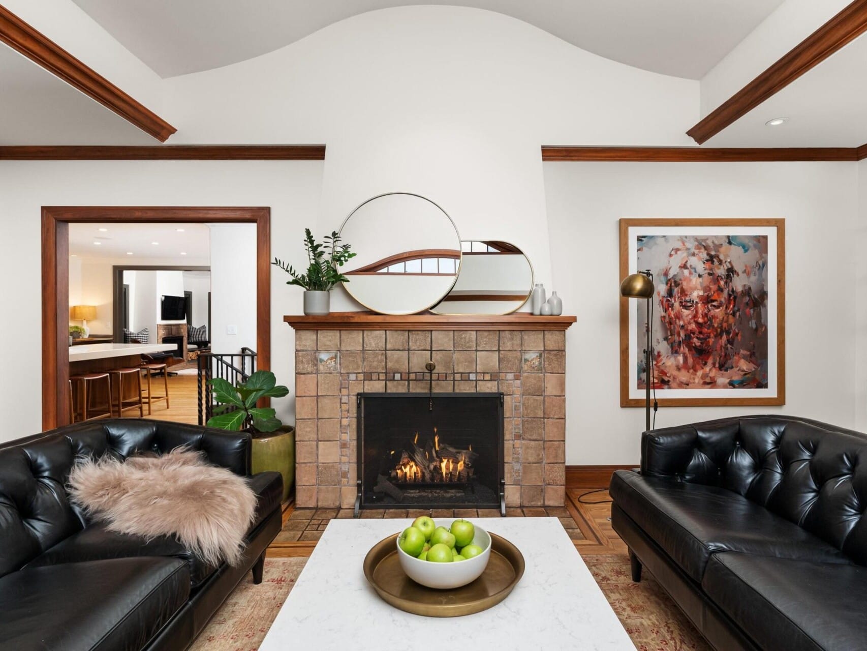 A cozy living room in Portland, Oregon real estate features a tiled fireplace flanked by black leather sofas. A round mirror and plants adorn above, while a bowl of green apples sits on the white coffee table. Artwork and wooden shelves with decor items complete this inviting space.