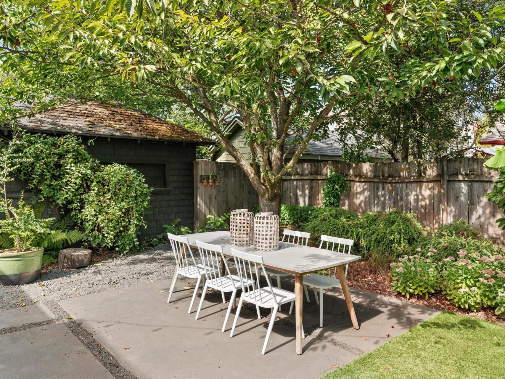 Outdoor patio with a white table and chairs under a large leafy tree, reminiscent of a serene Portland, Oregon garden. A wooden shed is in the background, surrounded by lush green plants and shrubs. The patio is part of a beautiful garden area with a well-maintained lawn.