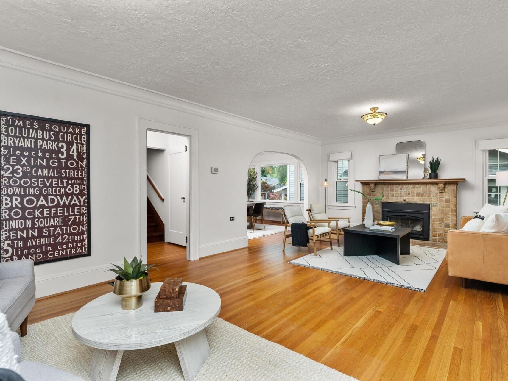 A cozy living room with hardwood flooring, a brick fireplace, and neutral-toned furniture. A white coffee table sits on a textured rug. A decorative sign hangs on the wall, and natural light filters through the windows, creating a warm ambiance.