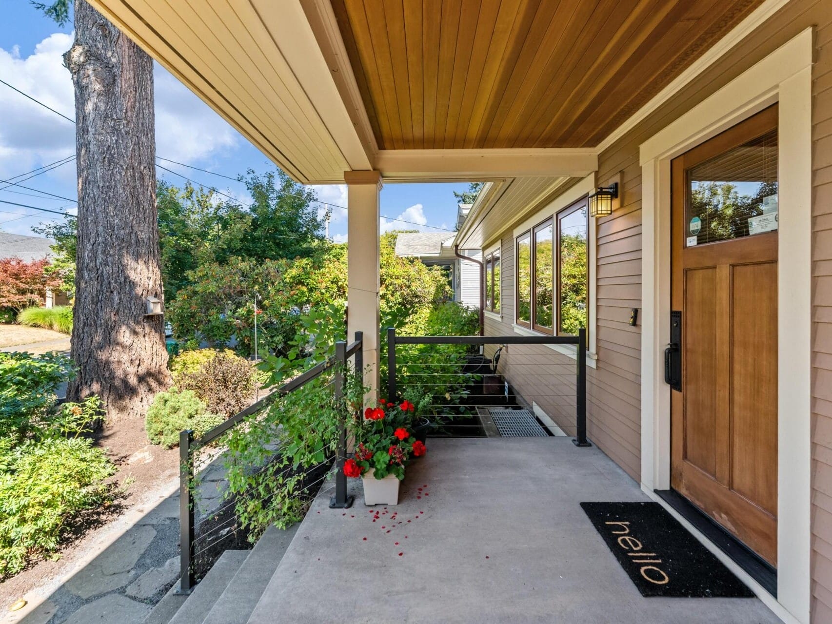 A cozy front porch in Portland, Oregon features a wooden ceiling and a welcome mat by the wooden door. Red flowers in a planter add color beside the steps, while lush greenery envelops the space on this sunny day.