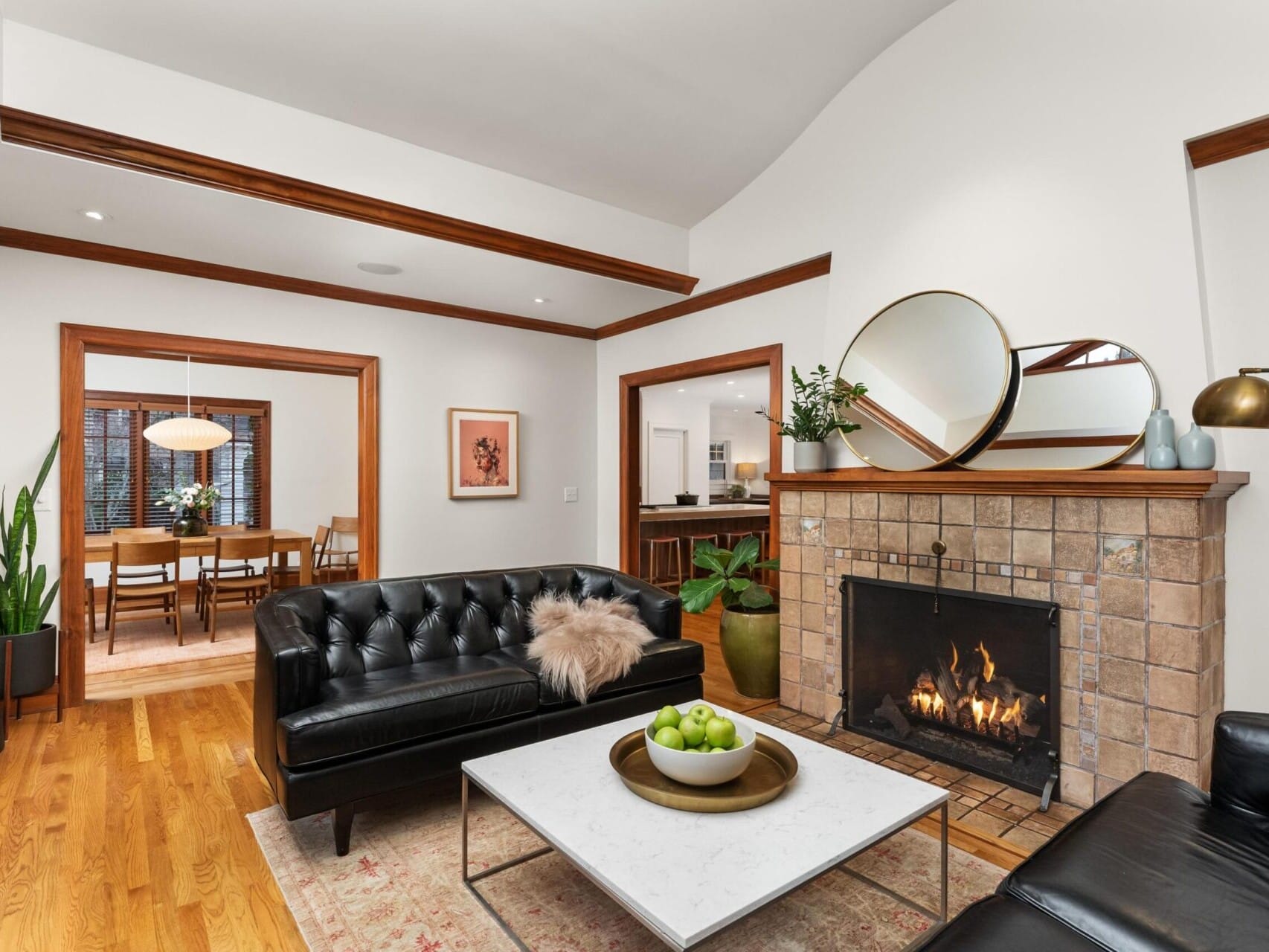 A cozy living room with a burning fireplace, black leather sofas, and a white marble coffee table holding green apples. A round mirror and plants adorn the mantel. Through the wooden archway, a dining area with a pendant light awaits, all exuding the charm of Portland Oregon real estate.