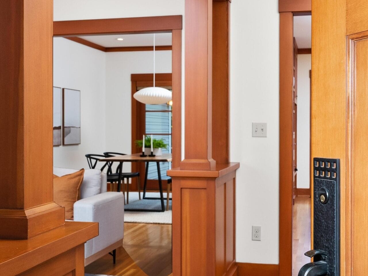 Open a wooden door to reveal a cozy Portland, Oregon interior with hardwood floors and warm wood trim. A modern dining area with a pendant light and wall art is visible in the background, along with part of a gray sofa. The space is well-lit and inviting.