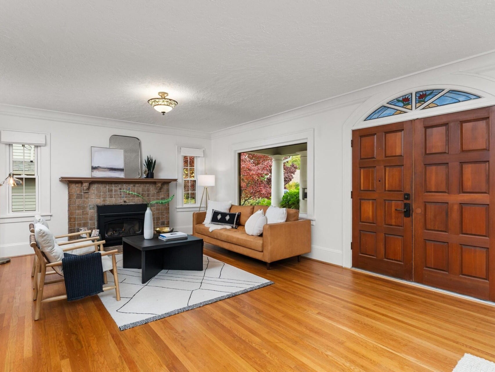 A cozy living room with a brown leather sofa, a dark coffee table, and a rug on wooden flooring. A brick fireplace is on one side, and a large wooden arched front door with stained glass panels is on the other. Large windows reveal leafy greenery outside.