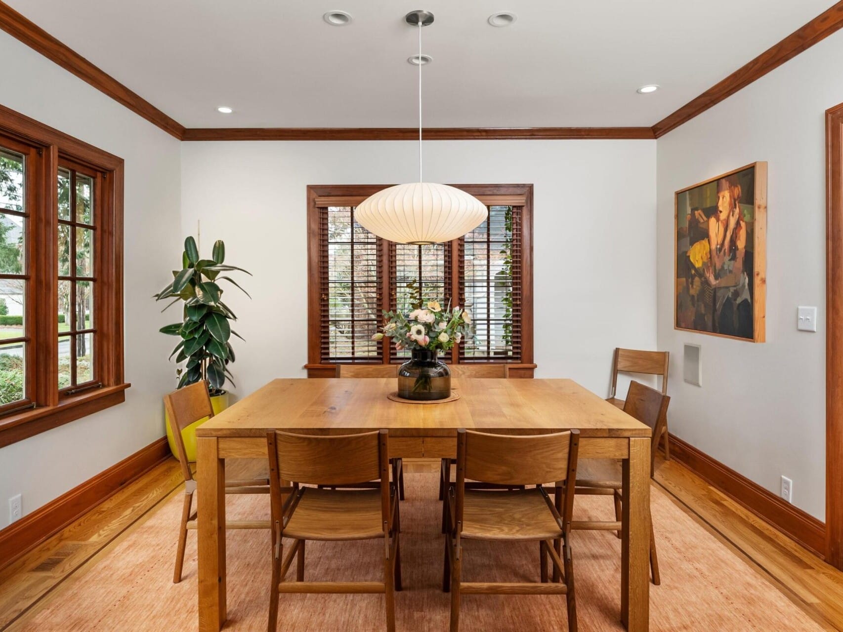 A dining room in a Portland Oregon Real Estate gem features a wooden table with six chairs on a beige rug. Above, a spherical light fixture casts a warm glow on the vase of flowers. Large windows grace the space, with a painting on the right and potted plant in the corner, showcasing elegance.